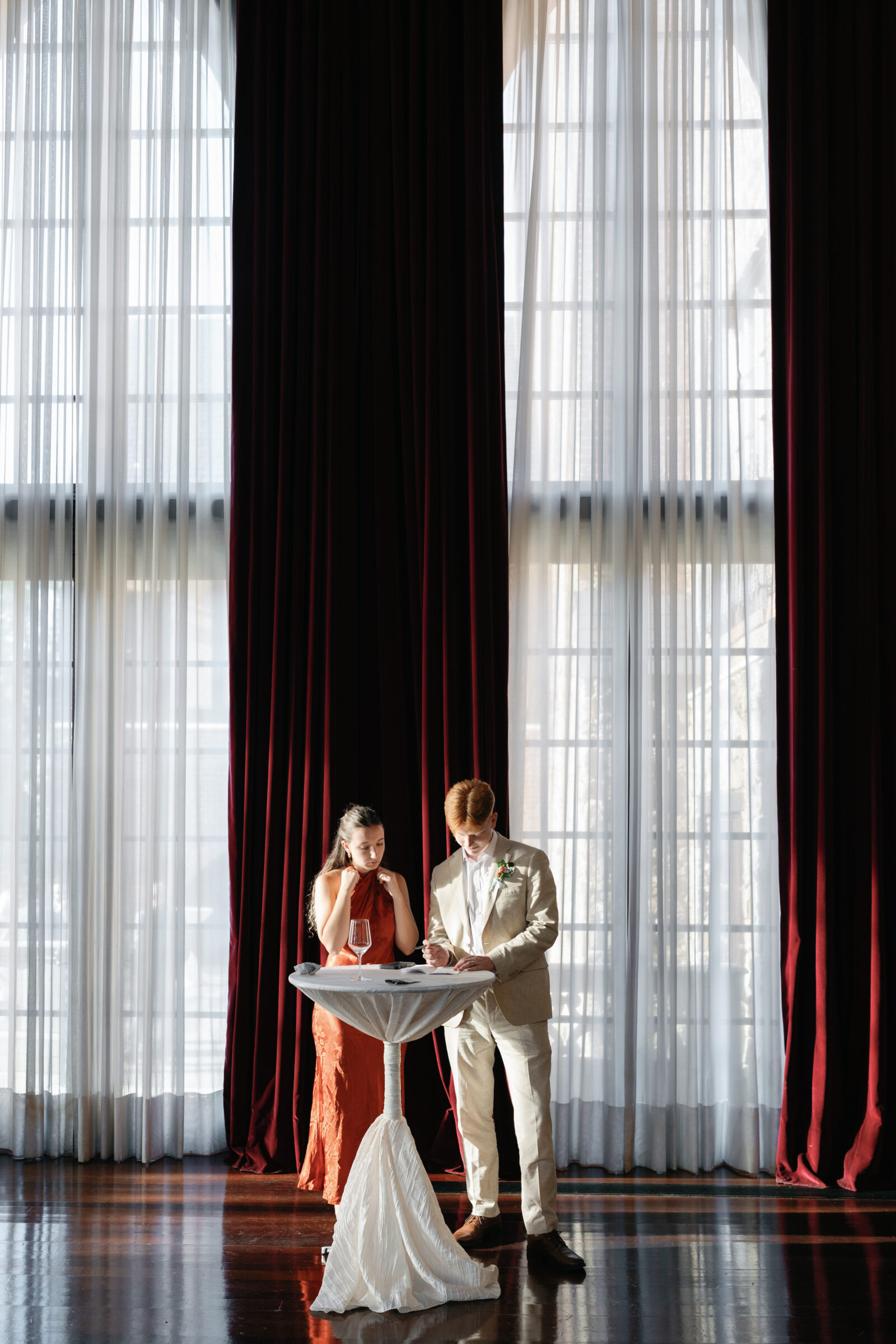 Wedding guests signing a keepsake during cocktail hour, framed by tall red velvet drapery and soft natural light — captured by a Richmond wedding photographer.