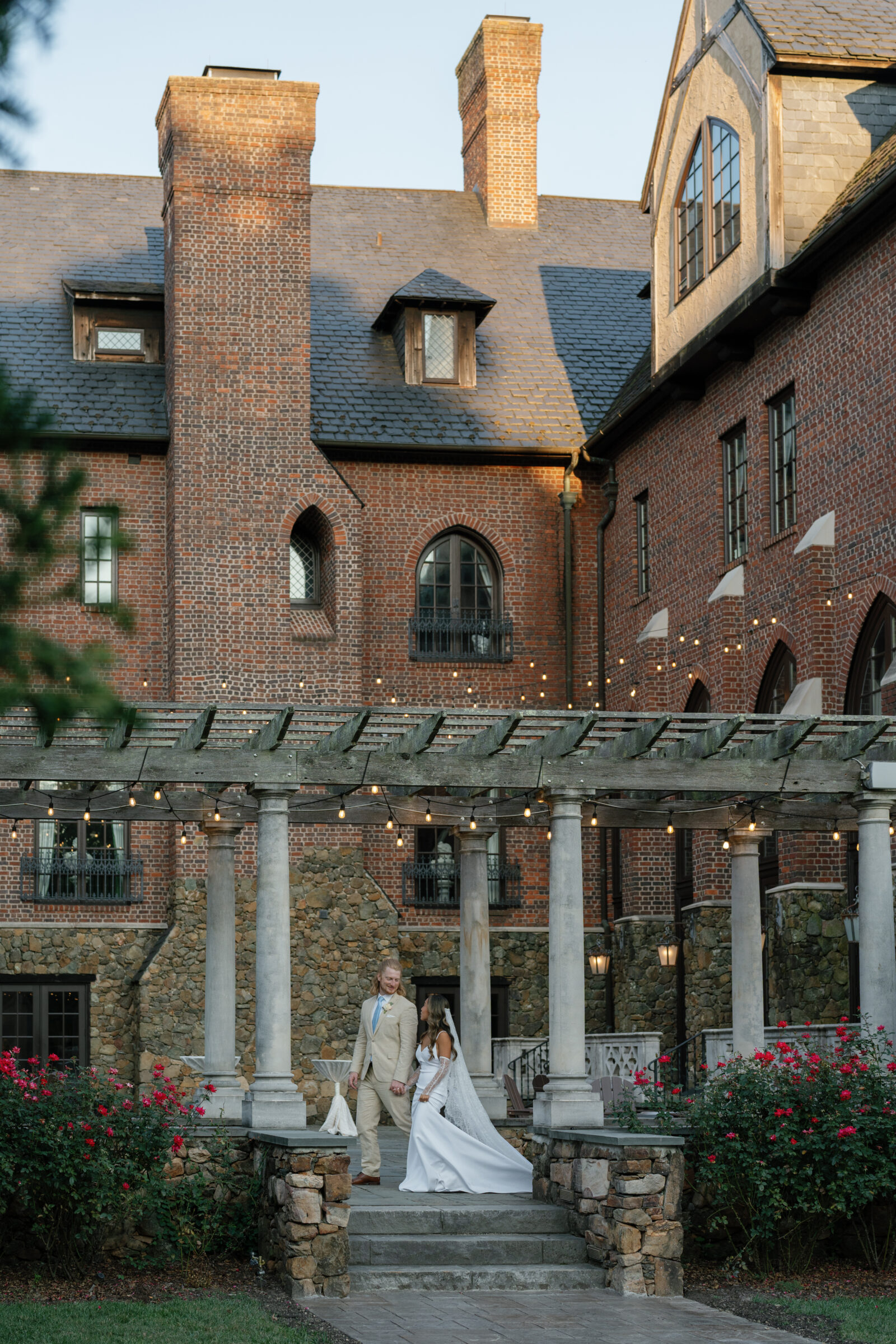 Bride and groom walking hand in hand beneath string lights and stone columns outside Dover Hall, framed by the estate’s brick architecture and garden roses — a romantic evening portrait captured by a Richmond wedding photographer during a Dover Hall wedding.