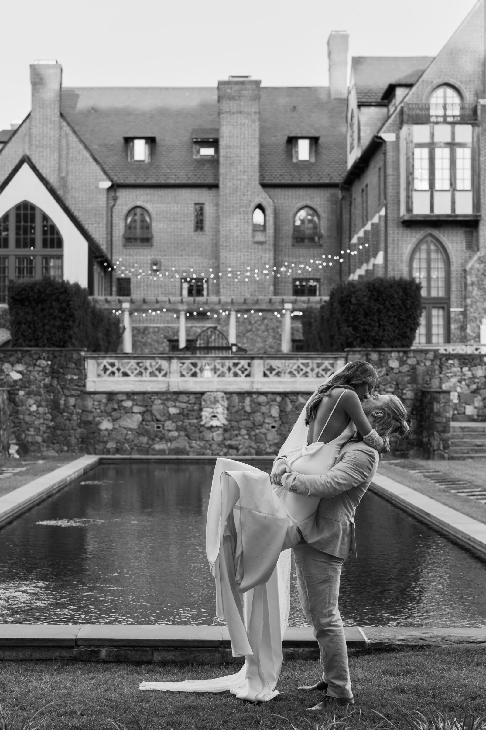 Groom lifting the bride beside the reflecting pool at Dover Hall, framed by string lights and the estate’s grand brick façade — captured by a Richmond wedding photographer during a Dover Hall wedding.