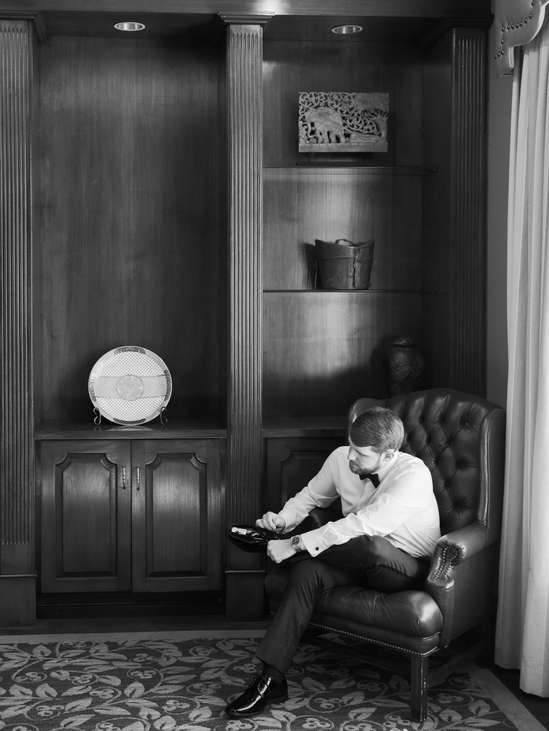 Hermitage Country Club Wedding | Groom sitting in a leather armchair tying his dress shoes in a wood-paneled room before the wedding.