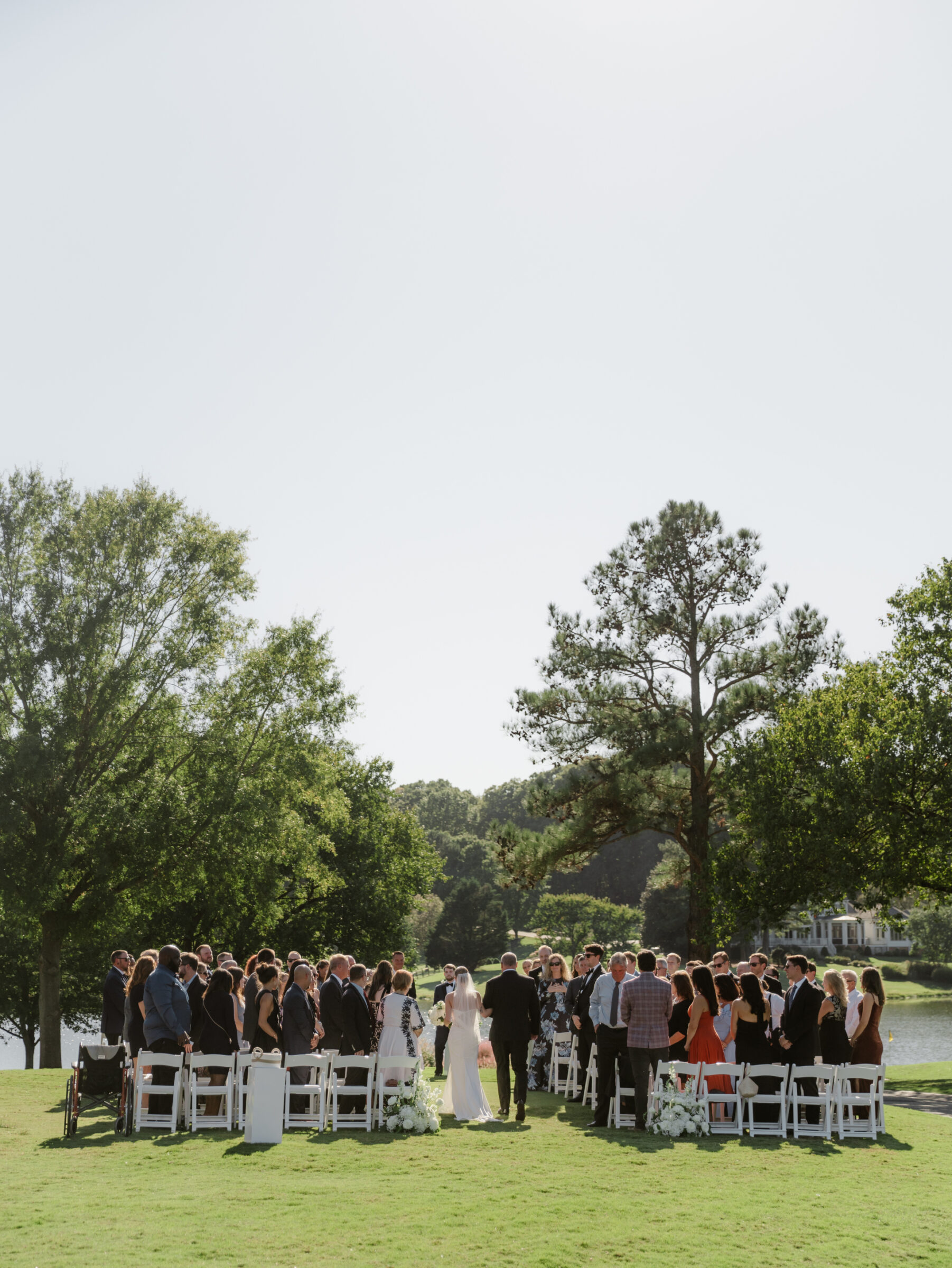 Hermitage Country Club Wedding | Outdoor wedding ceremony with guests standing as the bride walks down the aisle on a sunny day beside the water.