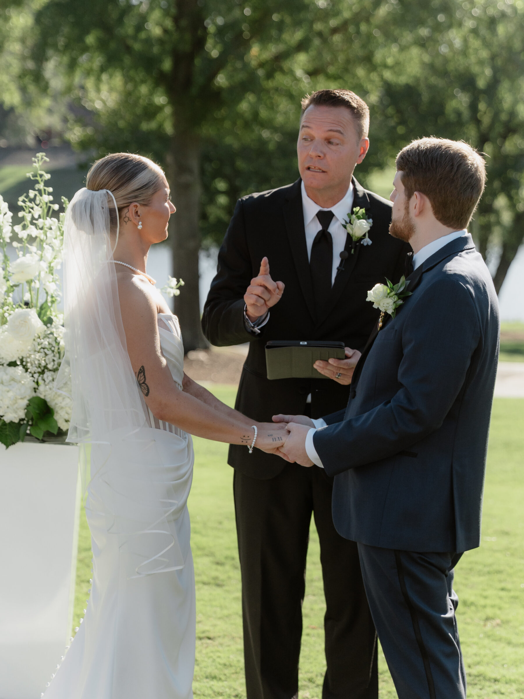 Bride and groom holding hands during their outdoor ceremony as the officiant speaks.