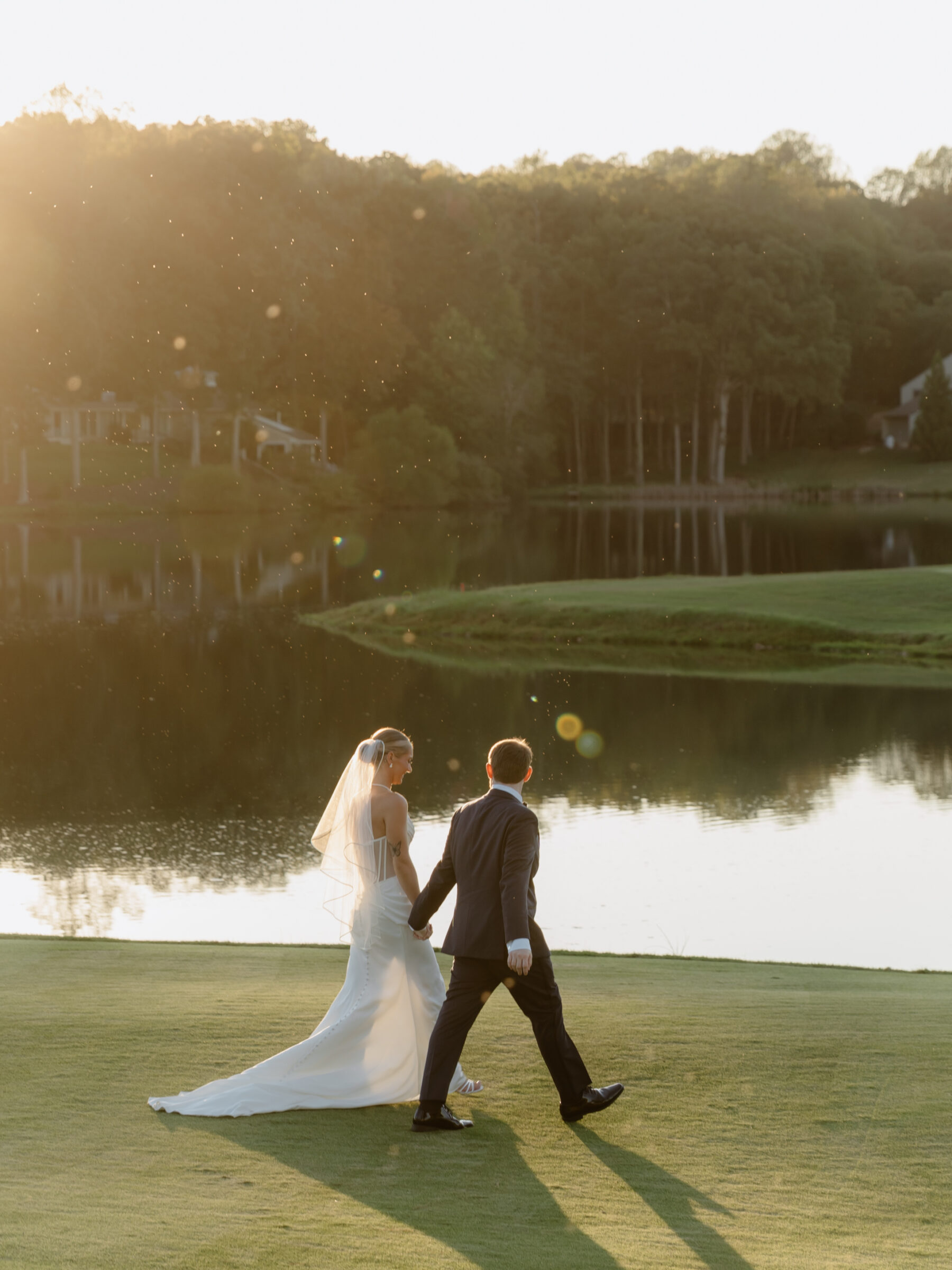 Bride and groom walking hand in hand along the water at sunset, with warm golden light surrounding them.