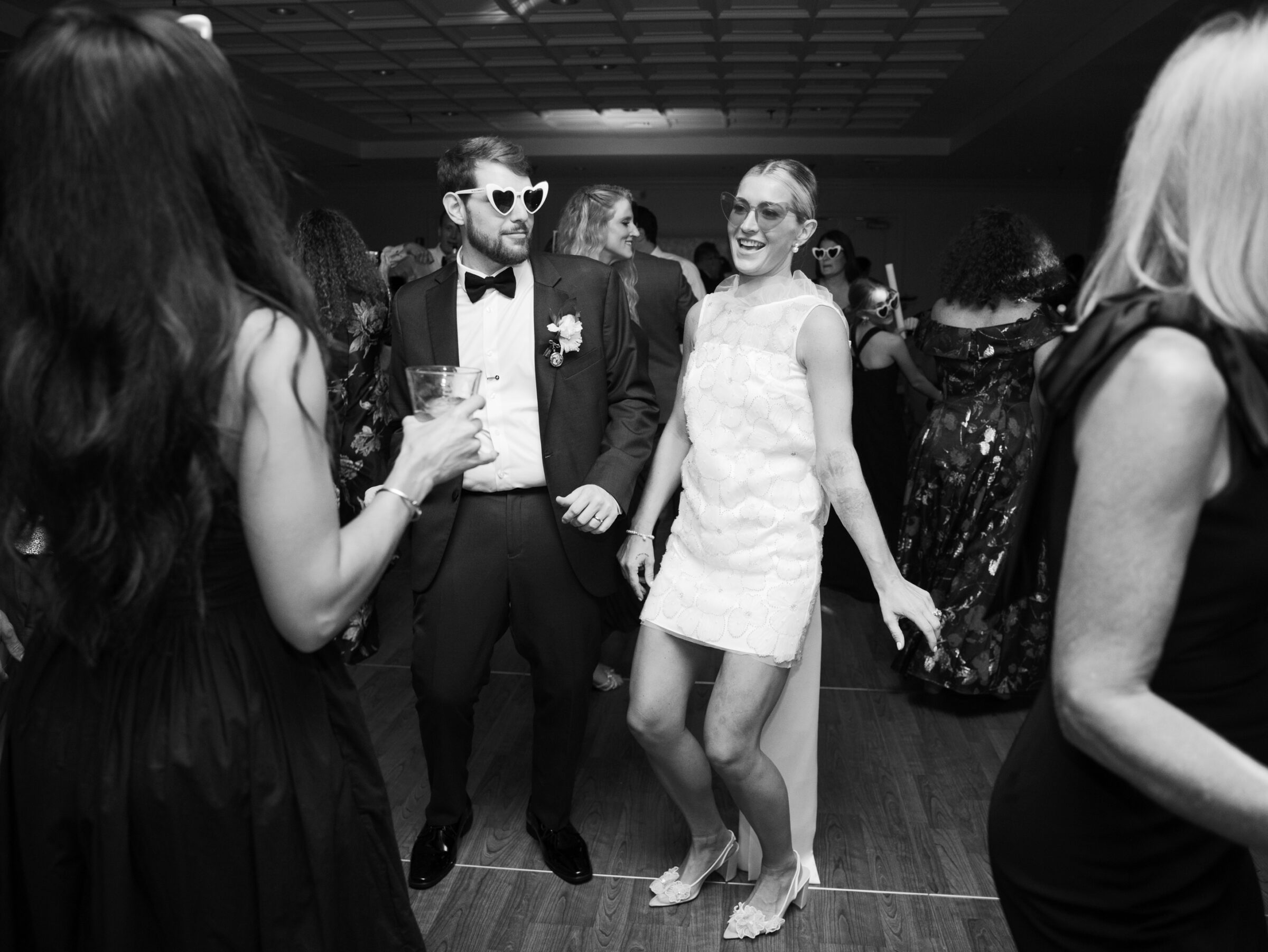 Bride and groom dancing at their reception while wearing fun party sunglasses, surrounded by guests on the dance floor.