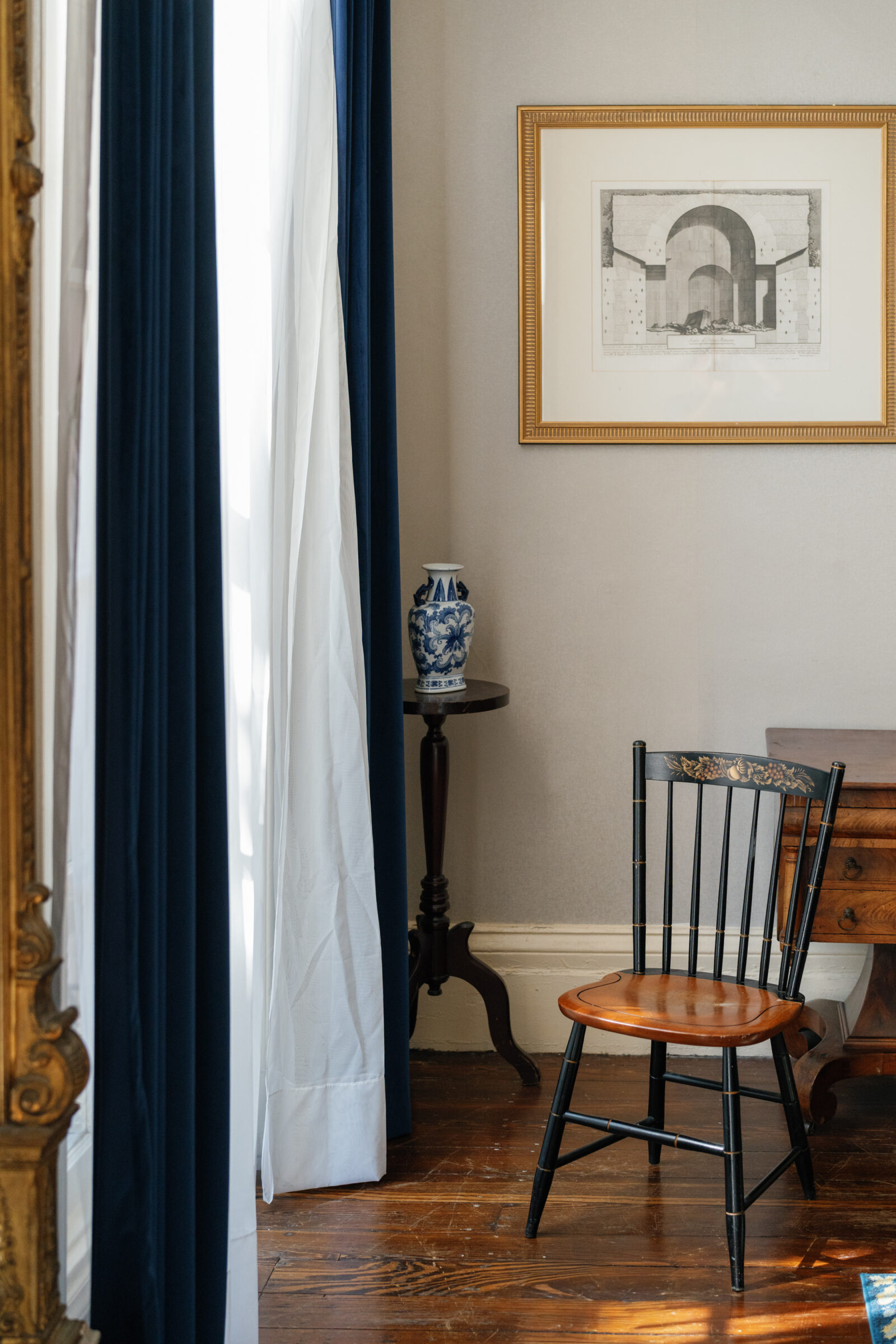 A sunlit corner inside Linden Row Inn with navy velvet curtains, a vintage wooden chair, a blue and white vase on a pedestal table, and a framed architectural print hanging on the wall.