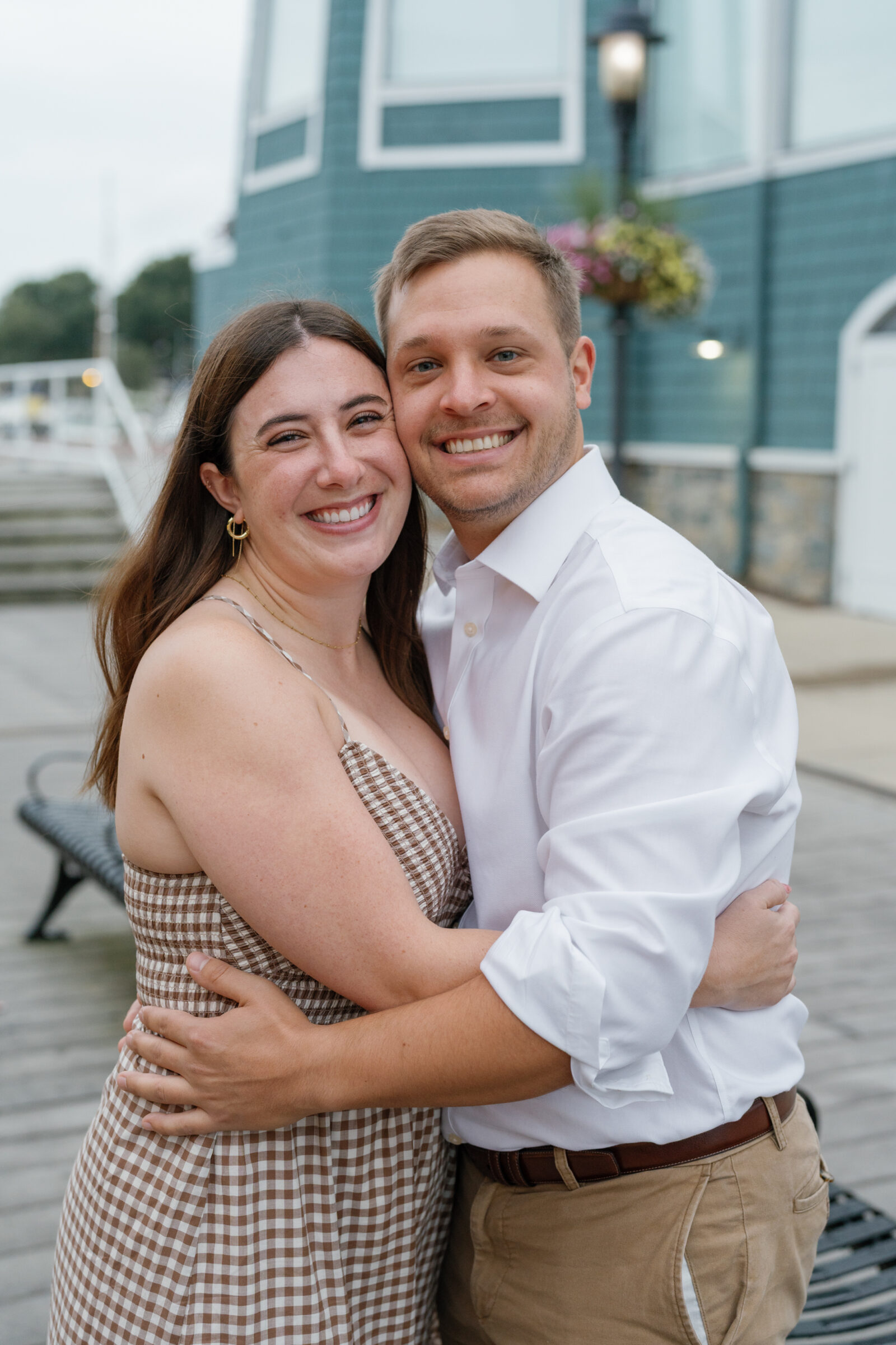Emma and Steven smiling and embracing by the waterfront during their Old Town Alexandria engagement photos, wearing a neutral gingham dress and white button-down for their sunrise session.