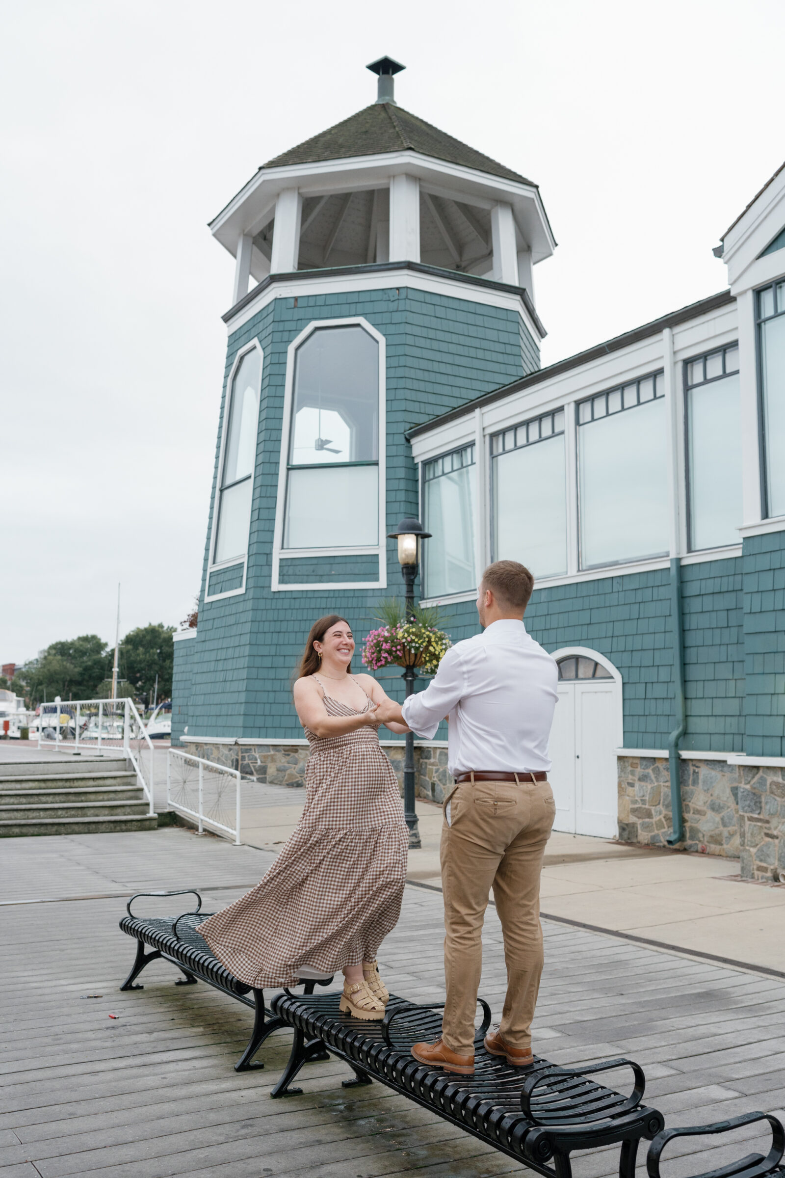 Couple dancing on a bench along the waterfront during their Old Town Alexandria engagement photos, with the Chart House building in the background on a breezy sunrise morning.