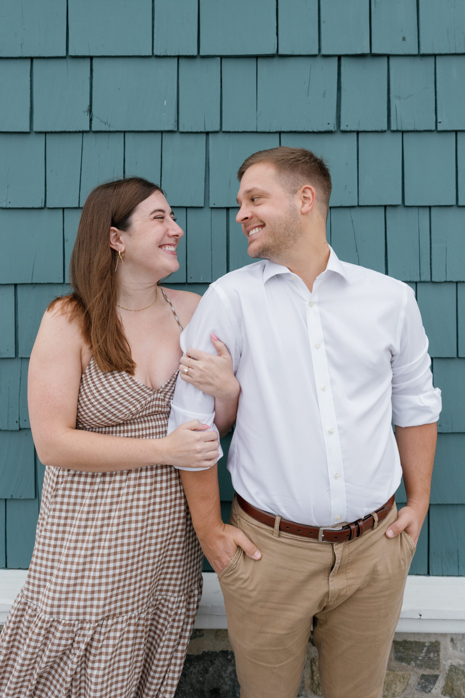 Couple smiling at each other while standing together along the waterfront in Alexandria, wearing a gingham dress and white button-down shirt.