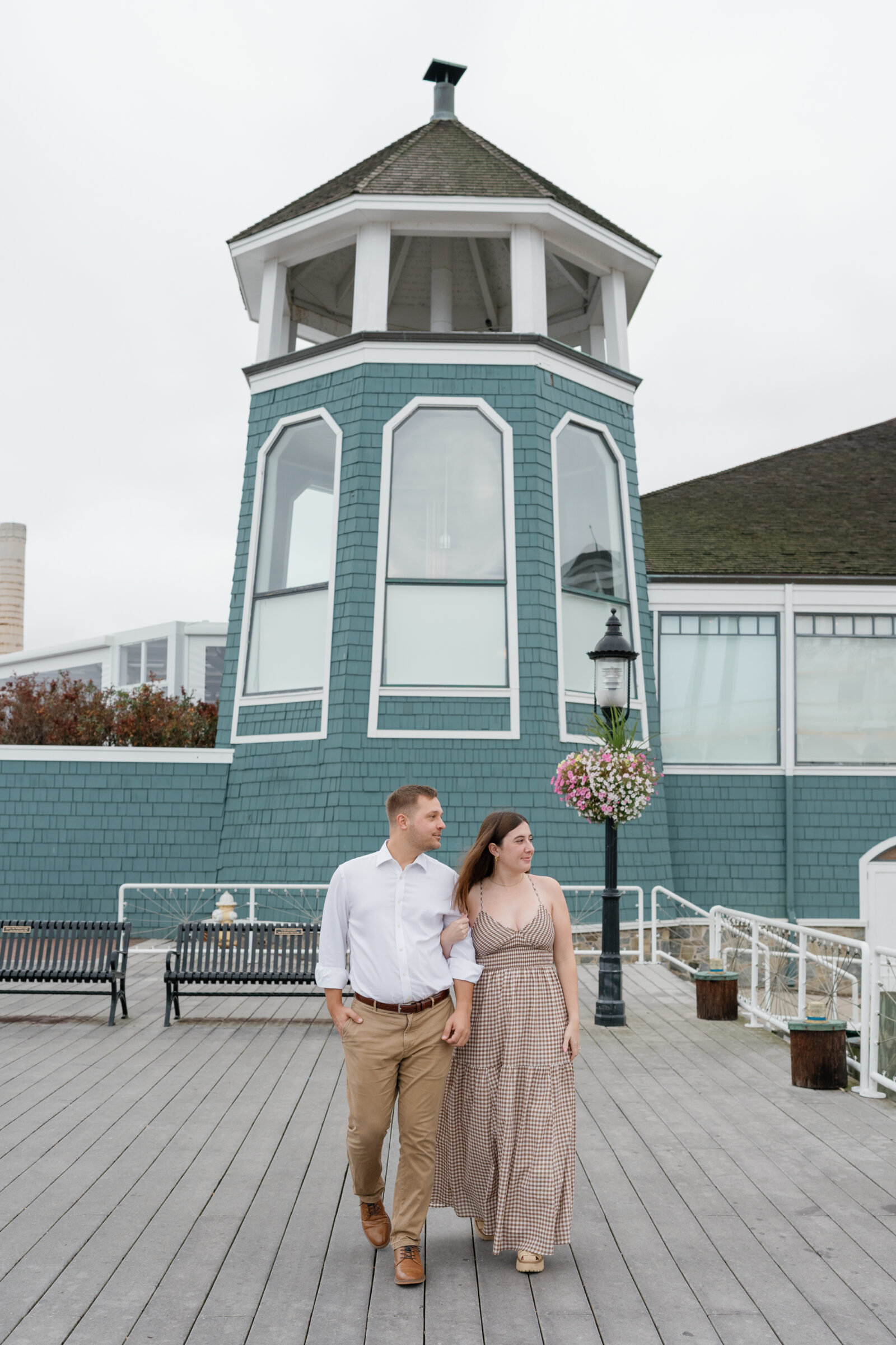 Couple walking arm in arm along the waterfront docks in Alexandria, wearing a gingham dress and white button-down shirt with the teal Chart House building behind them.