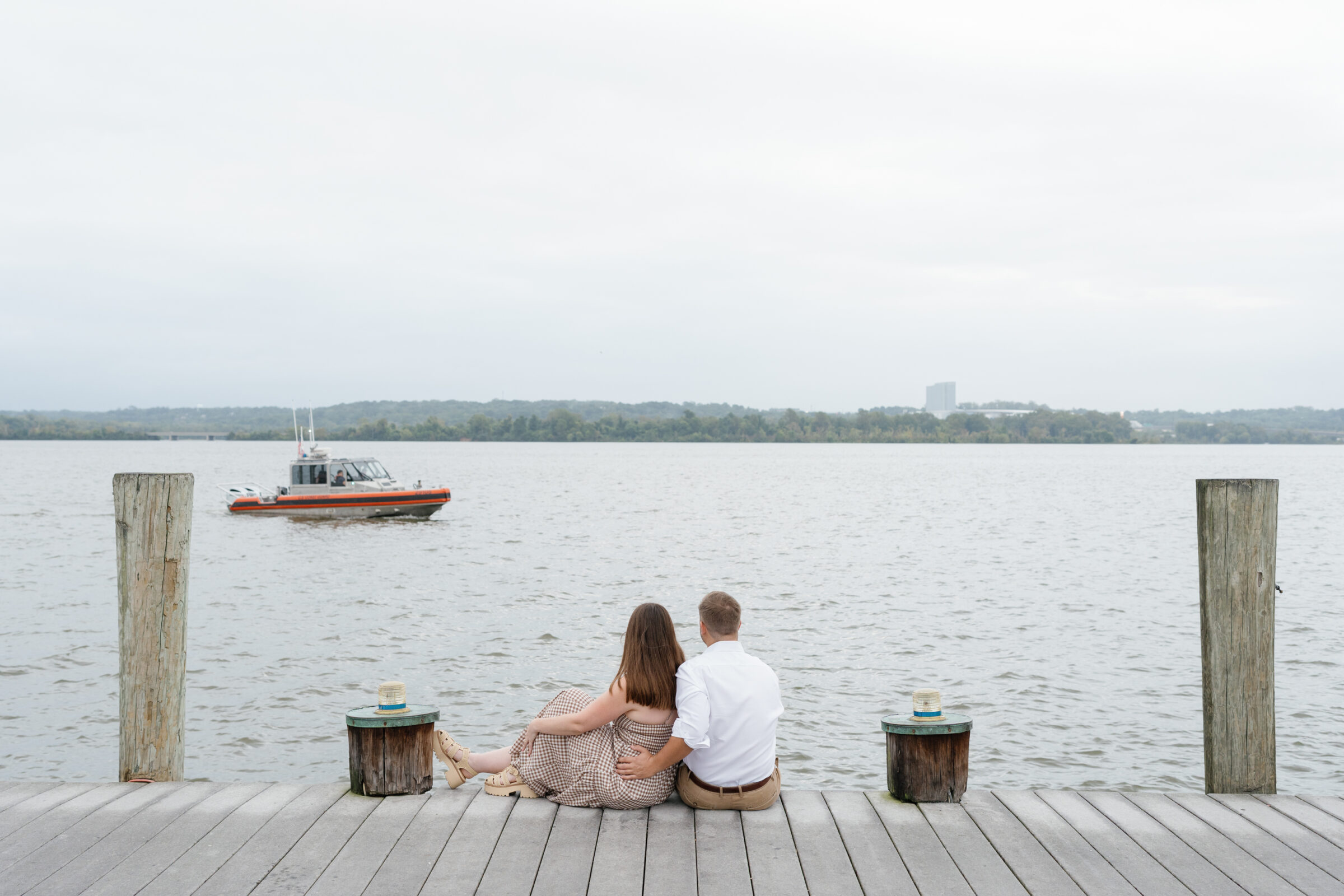 Old Town Alexandria Waterfront | Couple sitting together on the dock facing the water in Alexandria, with a boat passing on the Potomac River during their engagement session.