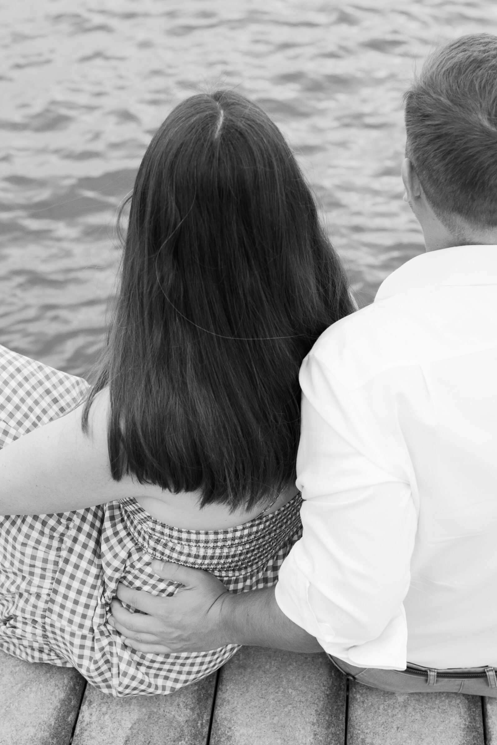 Close-up black and white photo of a couple sitting together on the dock, with his arm wrapped around her waist as they look out over the water.