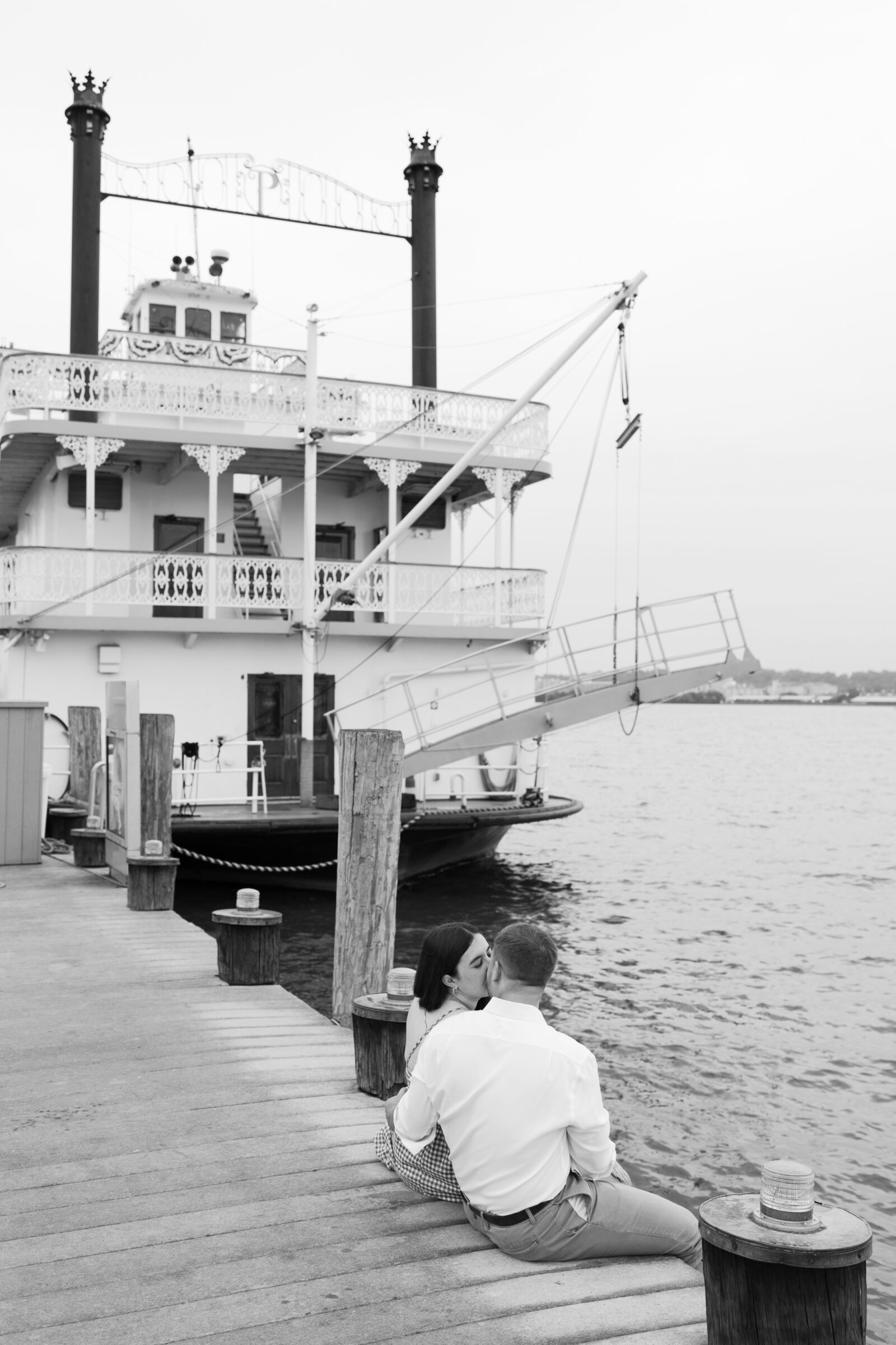 Black and white photo of a couple sharing a kiss while sitting on the dock near the riverboat in Alexandria, with the ornate paddle-wheel boat in the background.
