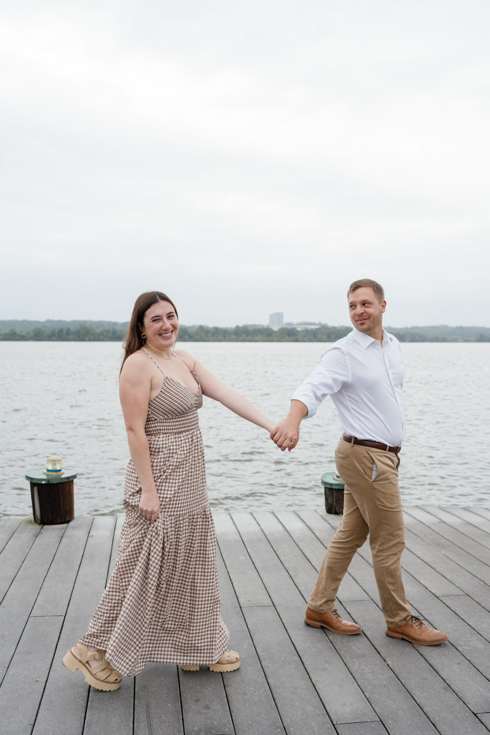 Couple holding hands and walking along the dock by the water during their engagement session, smiling at each other.