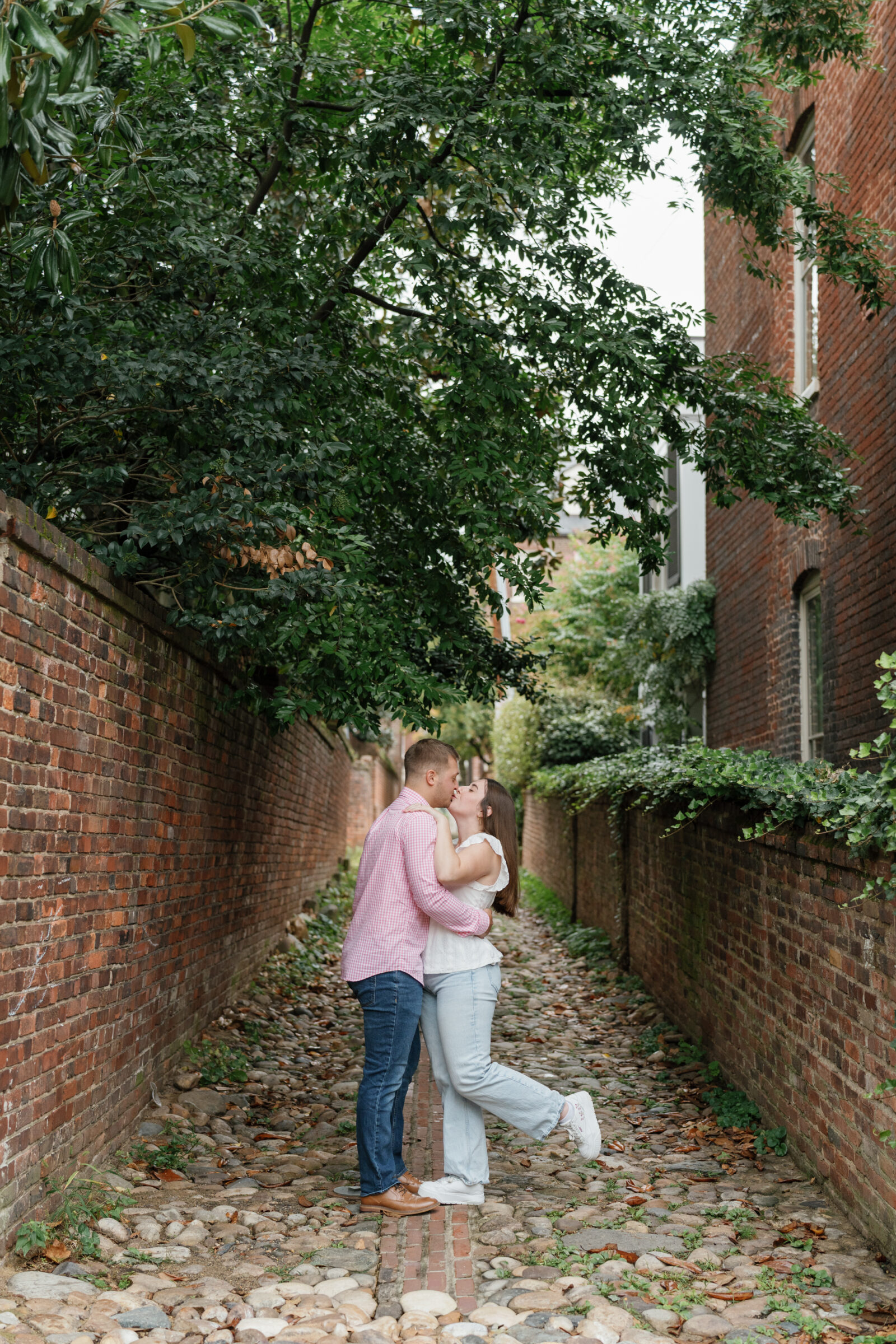 Couple kissing in a charming cobblestone alley in Old Town Alexandria, wearing casual outfits with jeans and a white blouse surrounded by historic brick walls and greenery.