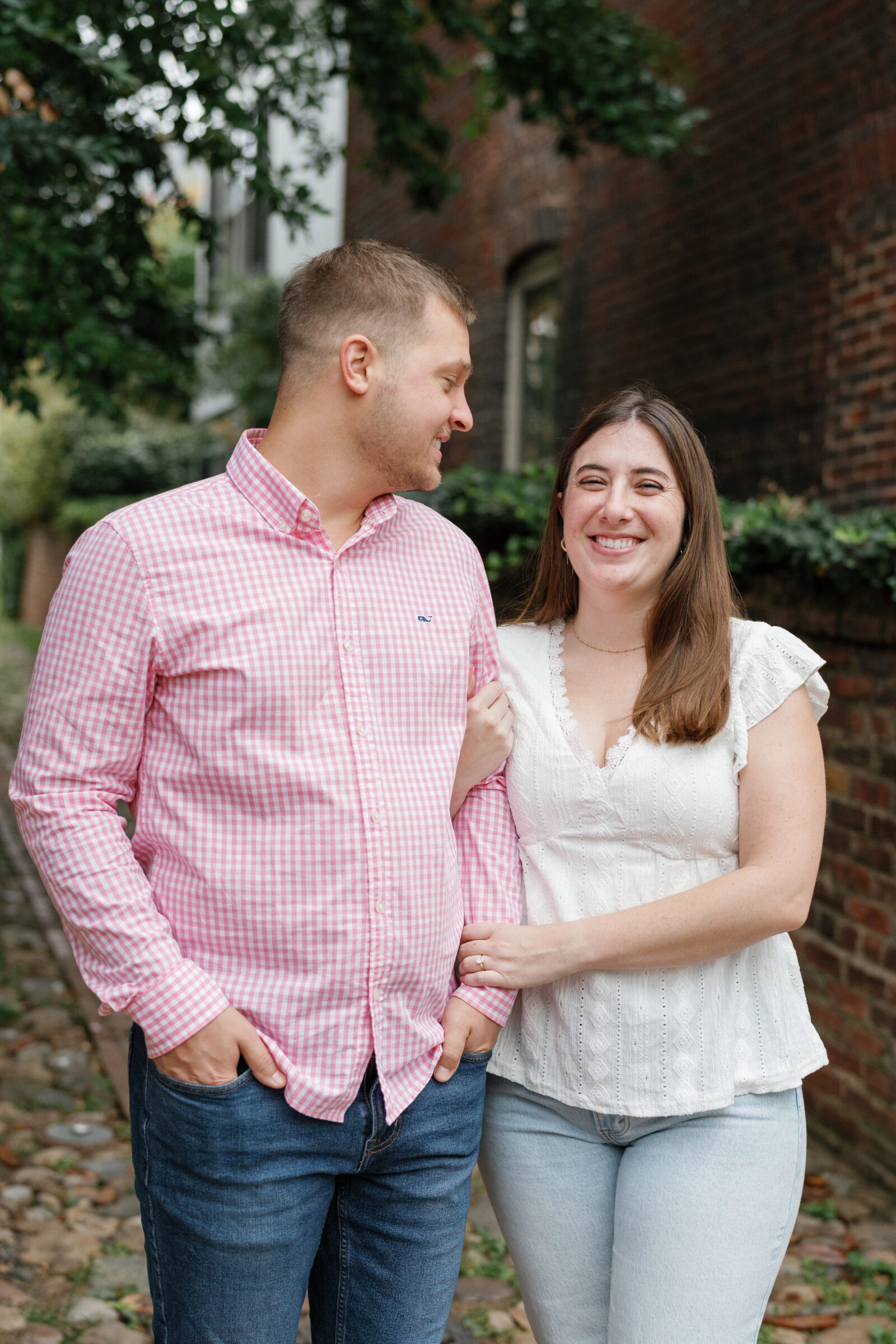 Old Town Alexandria Engagement Photos | Couple smiling together while standing arm in arm in a cobblestone alley, wearing casual jeans and a white blouse with a pink gingham shirt.