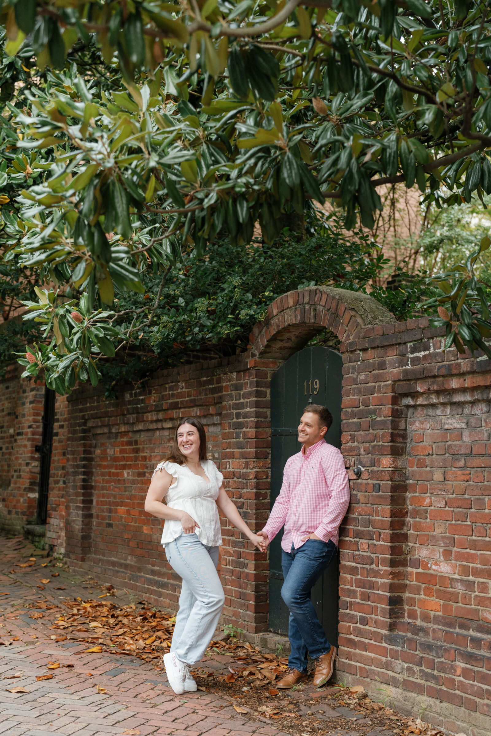 Couple holding hands and laughing together in front of a historic brick wall and arched green door, surrounded by greenery and fallen leaves.