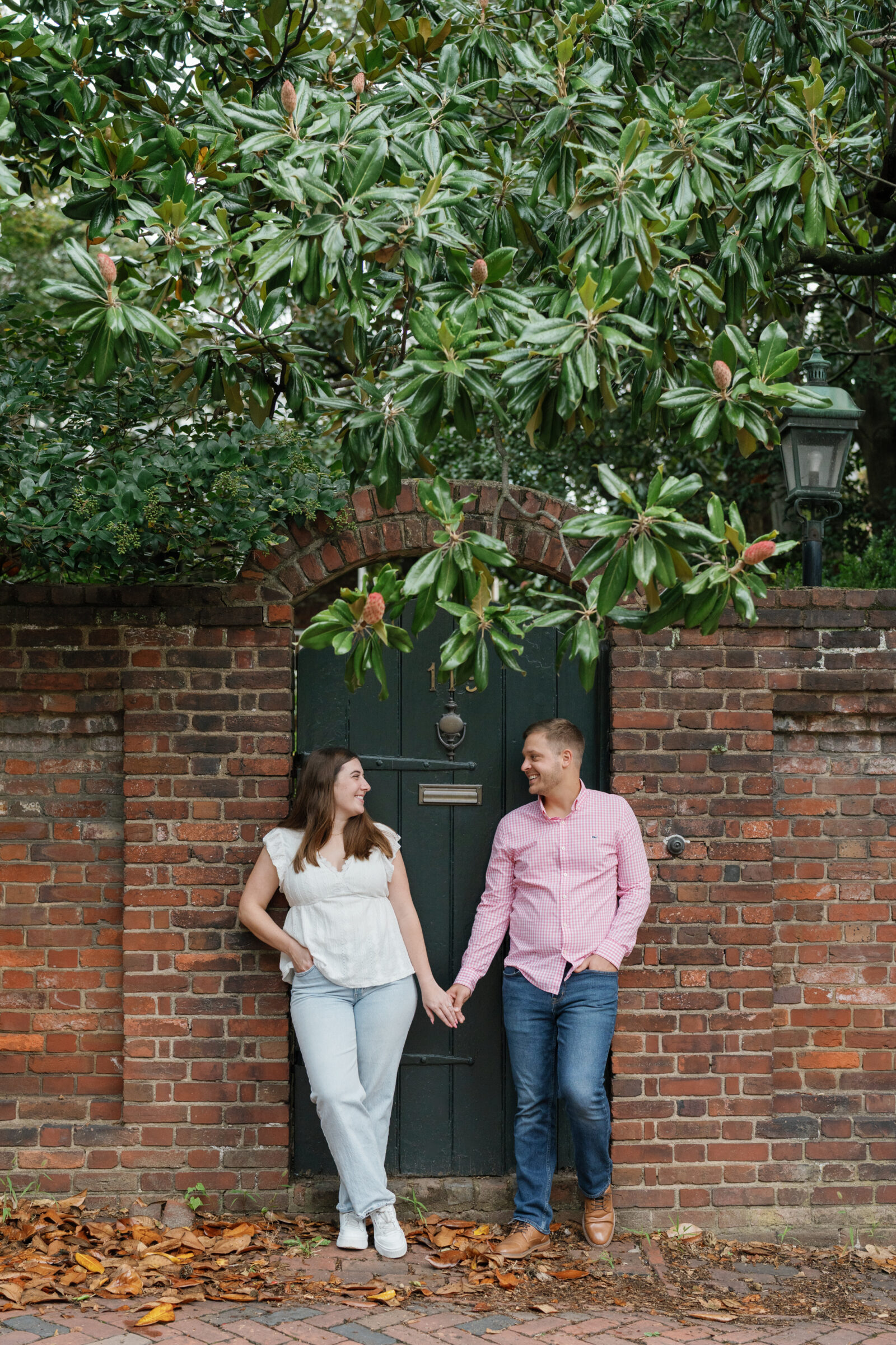 Couple holding hands and smiling at each other in front of a historic brick wall and dark green arched door in Old Town Alexandria, surrounded by magnolia branches overhead.