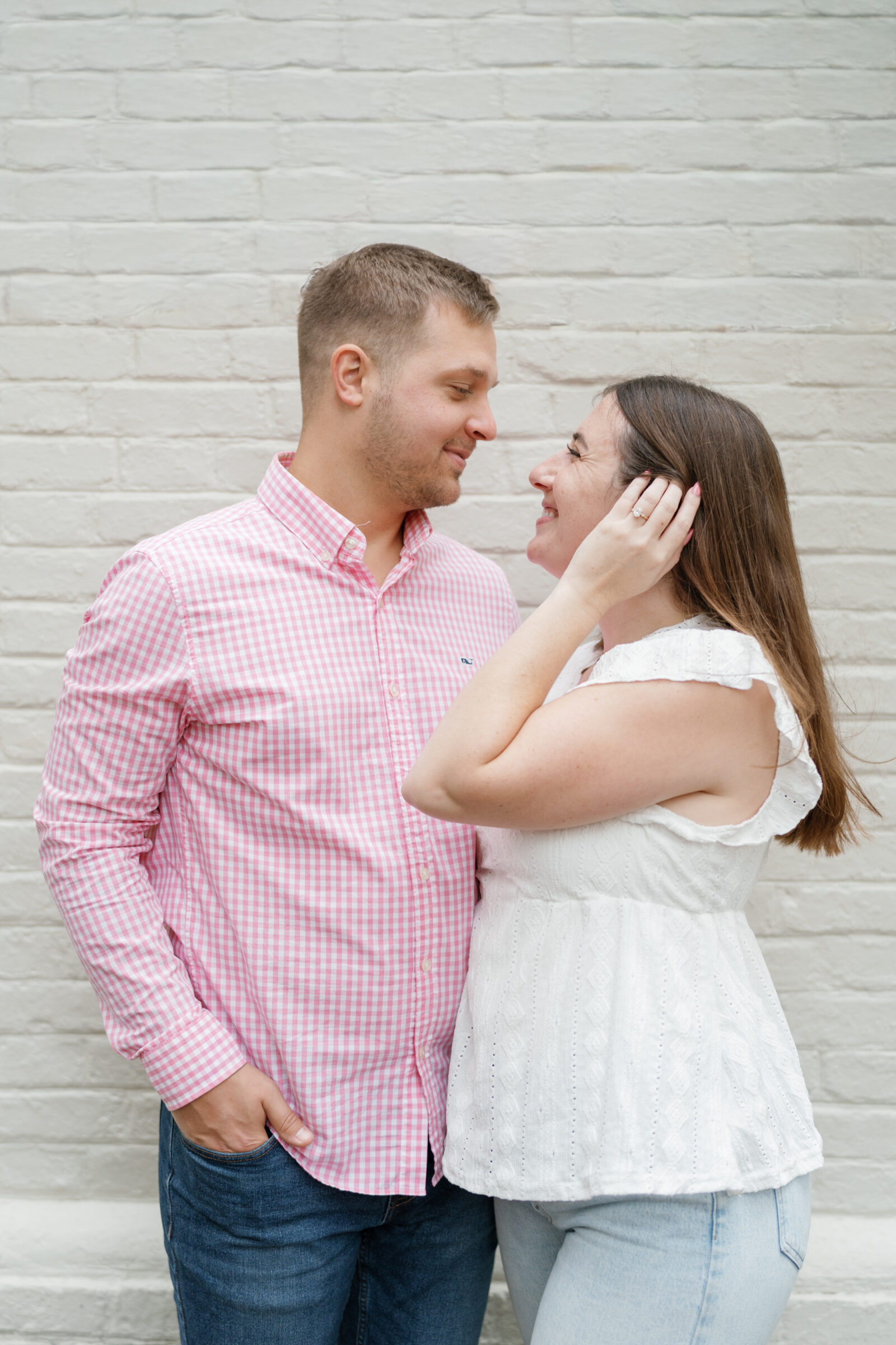 Couple smiling at each other up close, standing against a light brick wall during their engagement session.