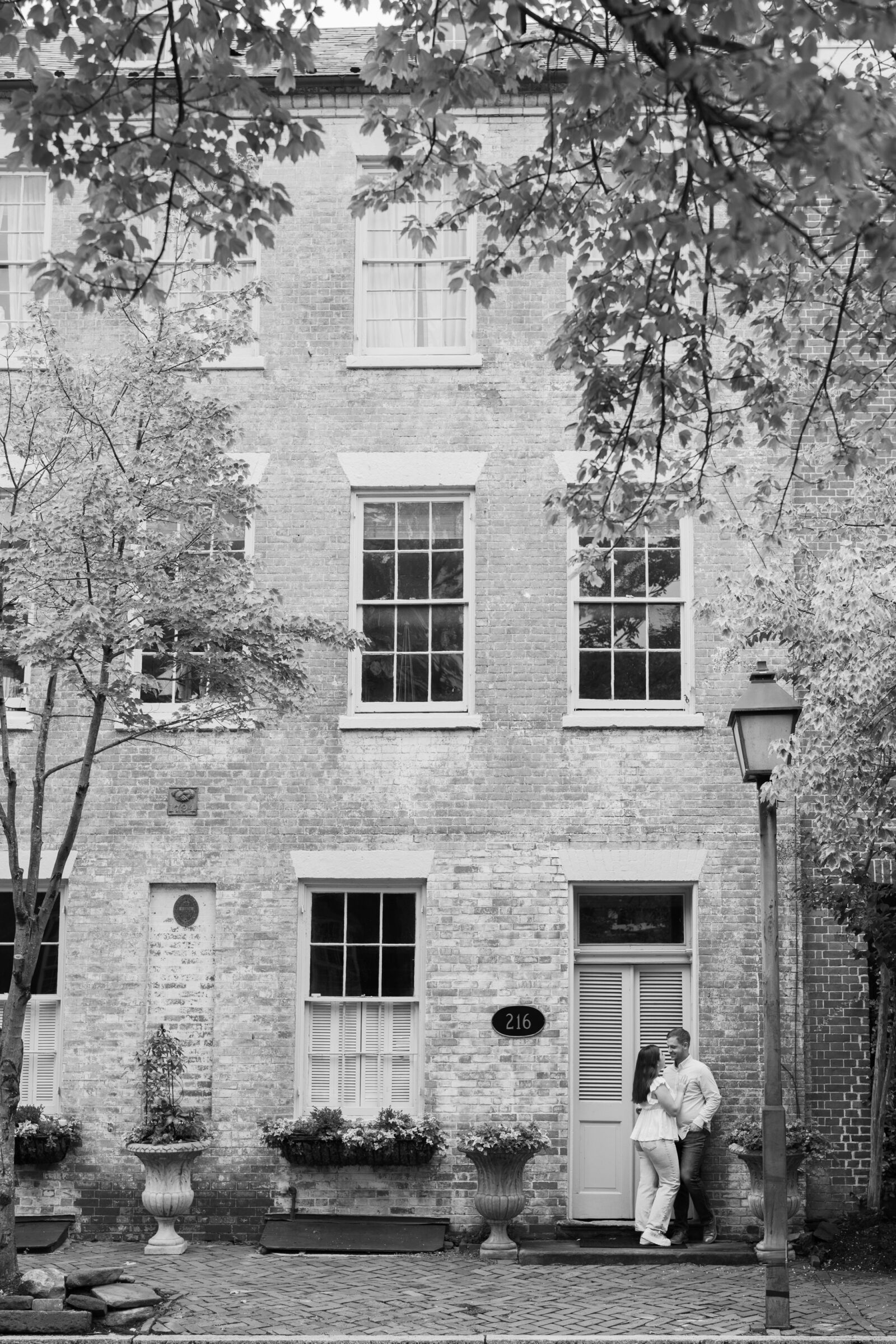 Couple standing close together by the front door of a historic brick townhouse lined with flower planters, framed by tree branches overhead.