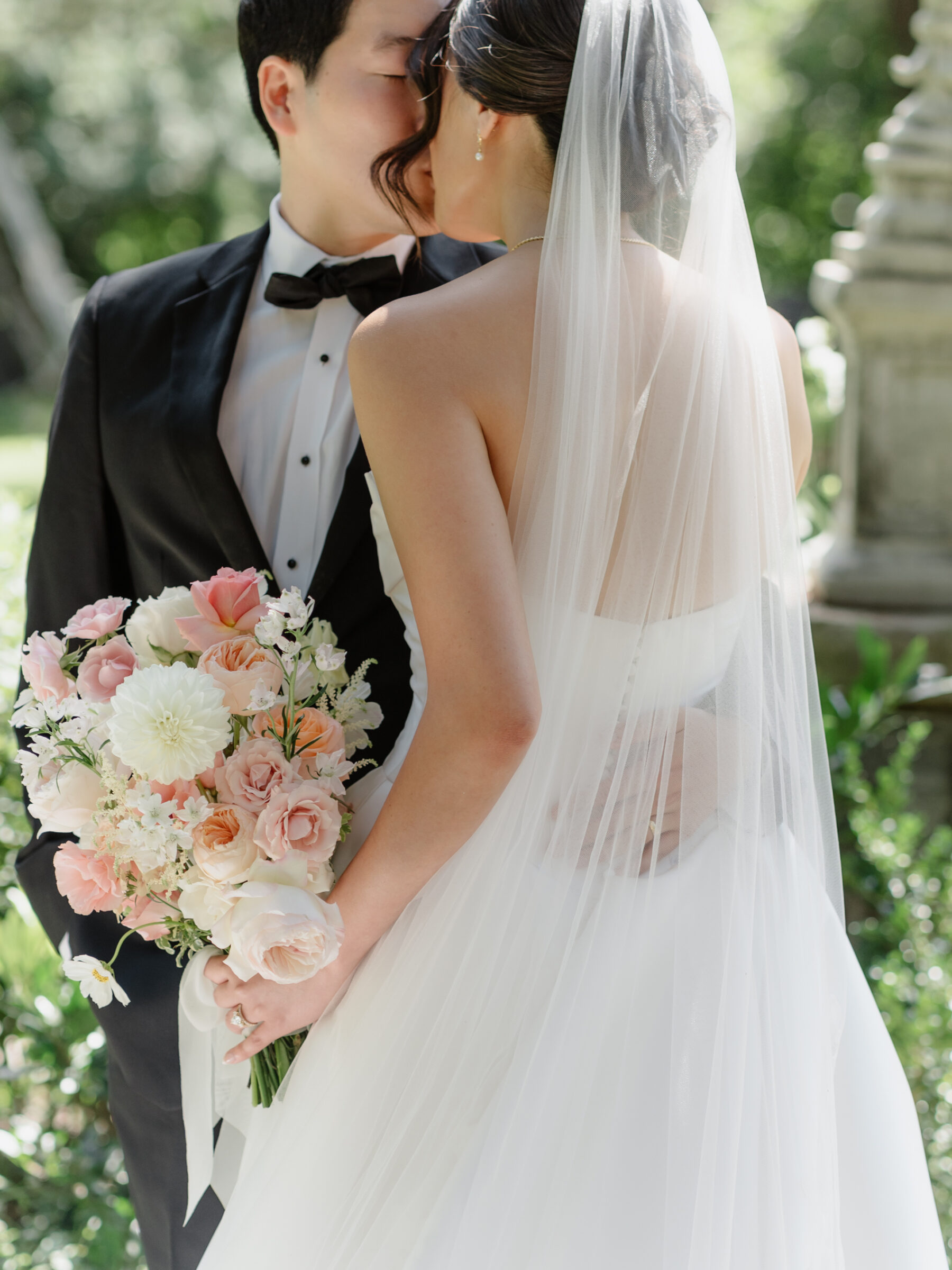 Rust Manor House Wedding | Close-up of the bride and groom sharing a kiss in the garden, with the bride holding a soft pink and white bouquet and her veil draped over them in the sunlight.