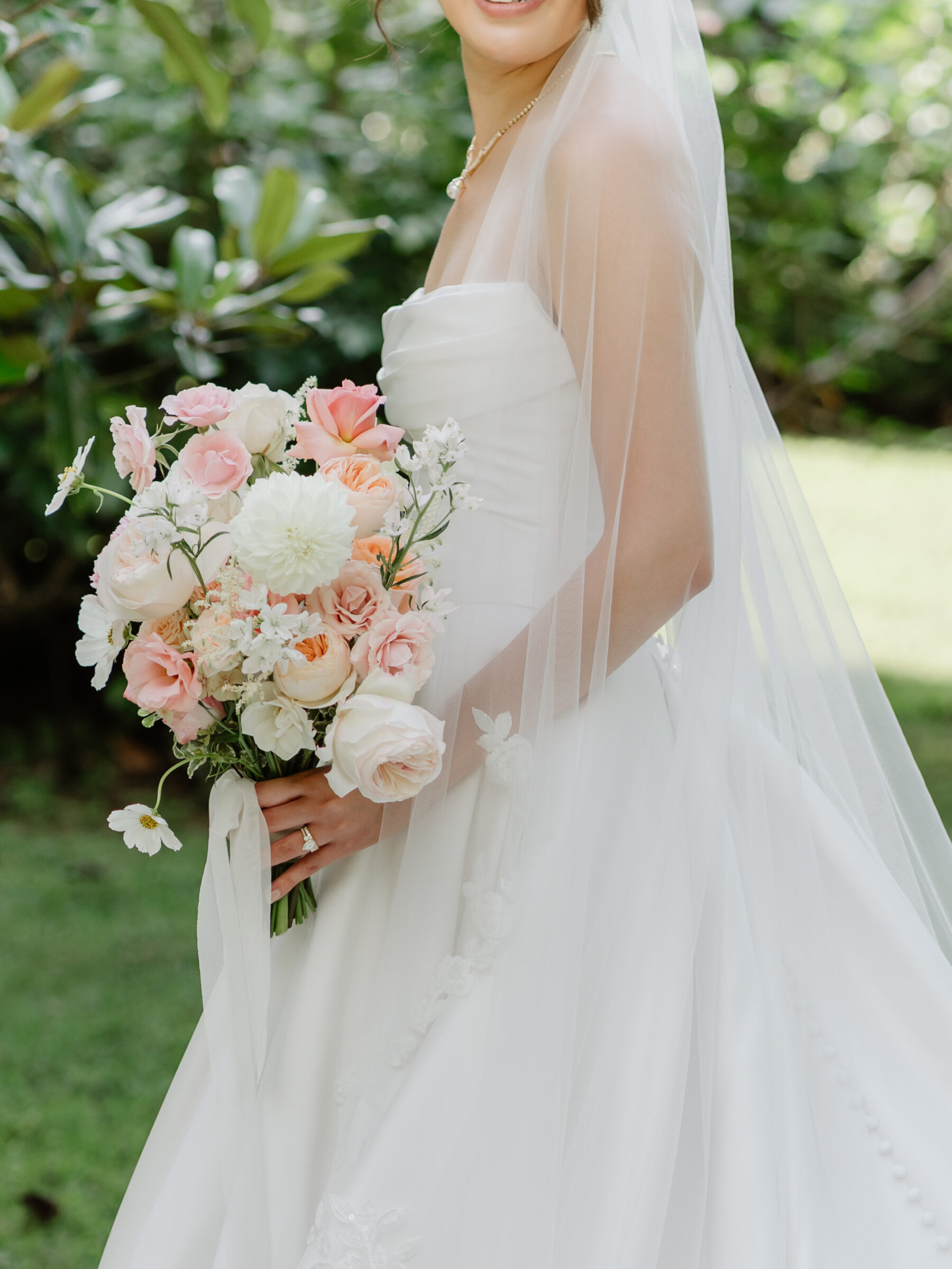 Bride holding a soft pink and white bouquet, with her veil and timeless gown flowing in the garden light.