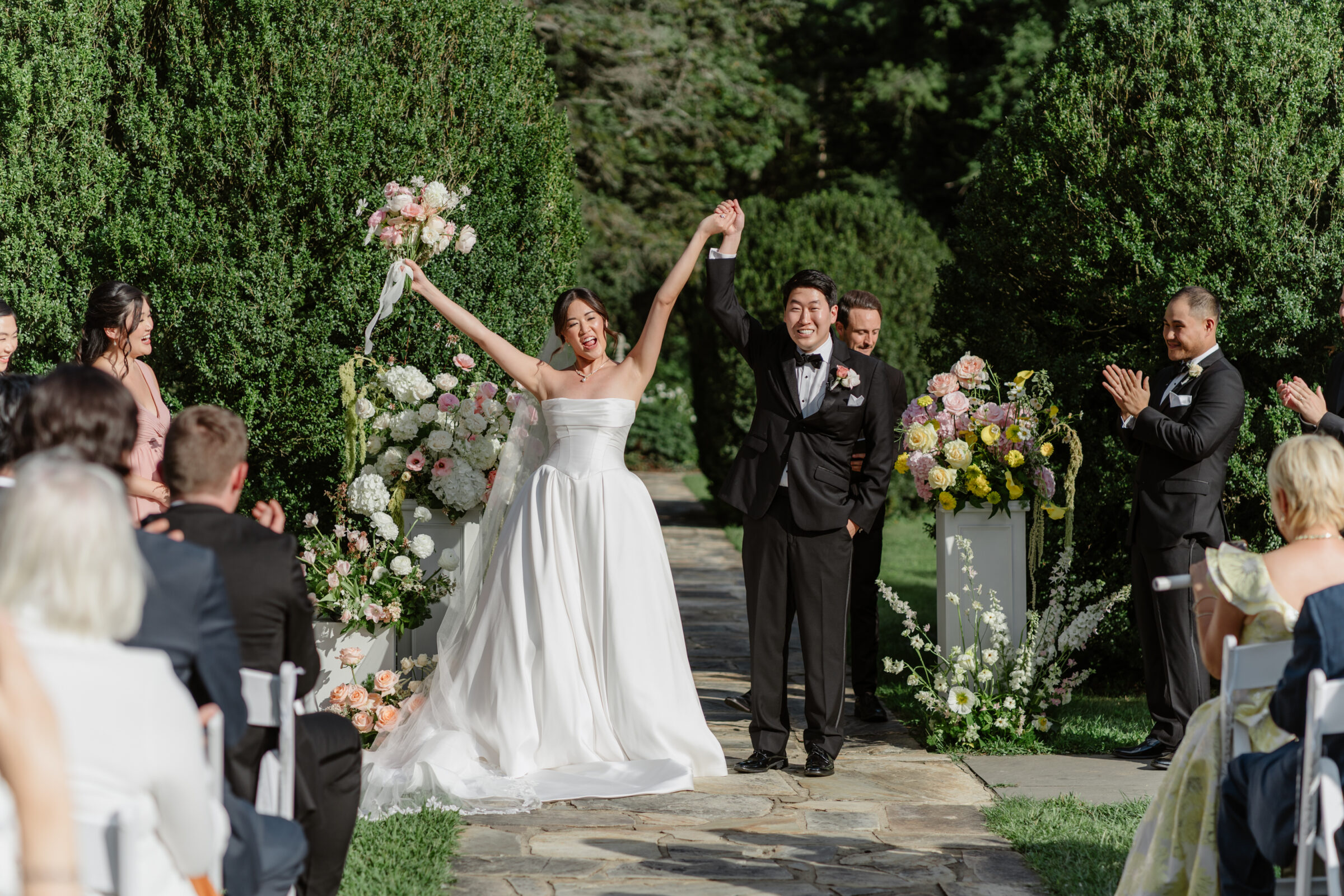 Rust Manor House Wedding | Bride and groom celebrating at the end of their garden ceremony, holding hands triumphantly as guests applaud and floral arrangements frame the aisle.