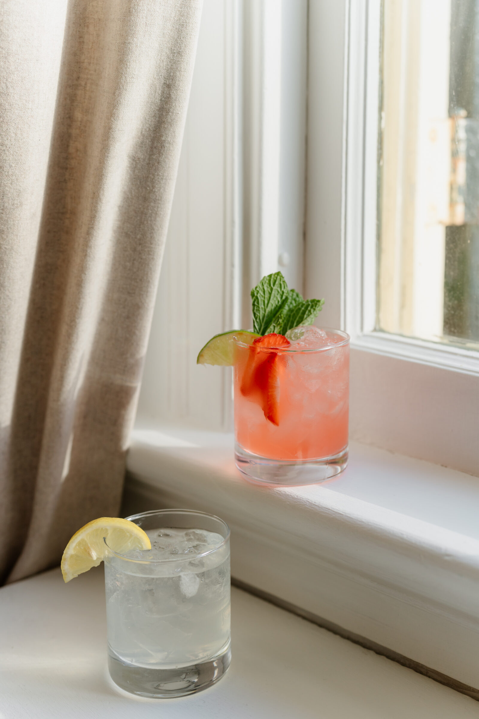 Two signature cocktails sitting on a sunlit windowsill, including a pink drink garnished with mint, lime, and strawberries, and a citrus cocktail with a lemon slice.