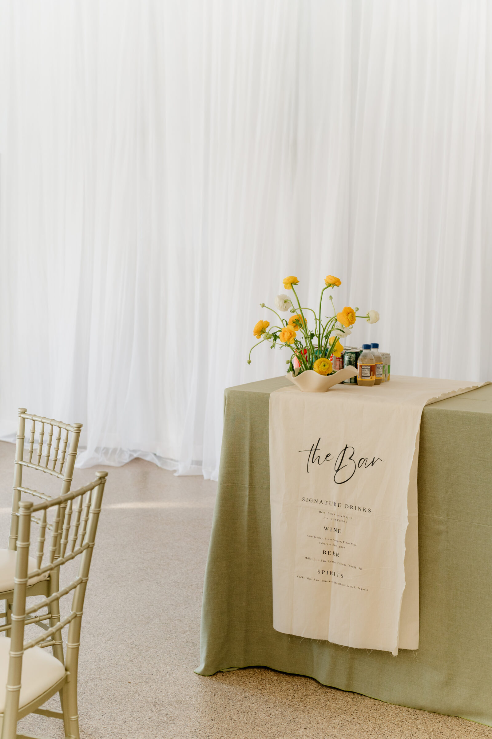 Wedding bar setup with a sage linen table, a handwritten bar menu runner, and a small arrangement of yellow ranunculus against soft white draping.
