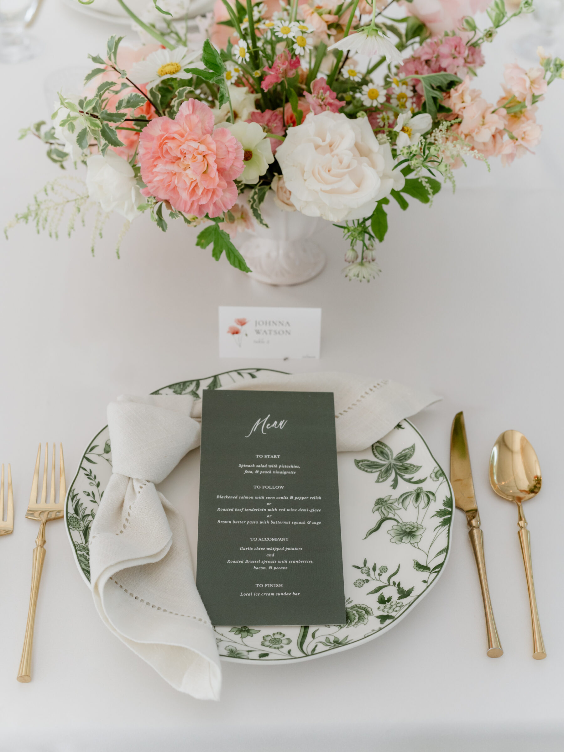 Rust Manor House Wedding reception place setting with a green printed menu on floral china, gold flatware, and a soft pink and white centerpiece.