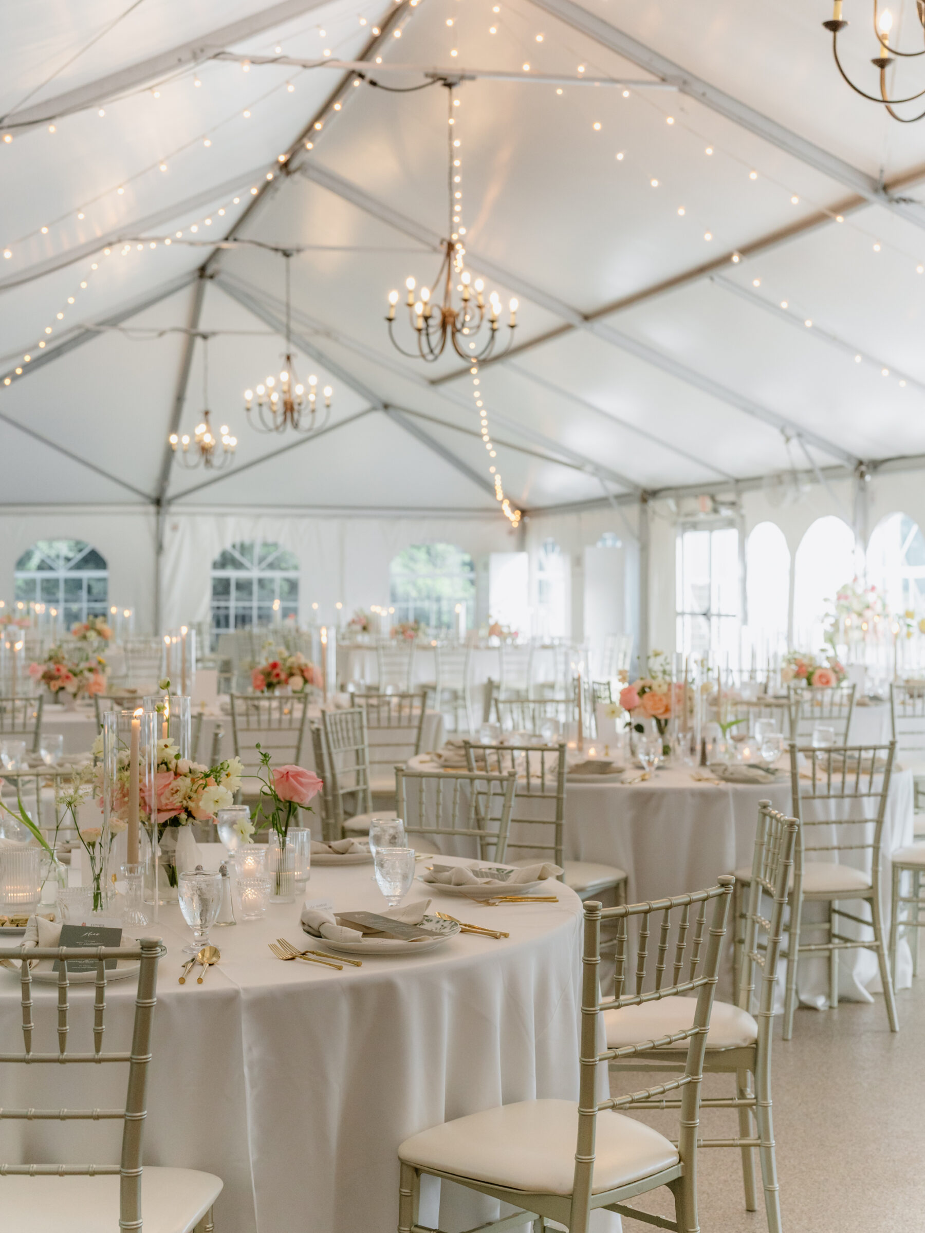 Tented Rust Manor House wedding reception with soft draping, chandeliers, and round tables set with elegant floral centerpieces, candles, and gold-accented place settings.