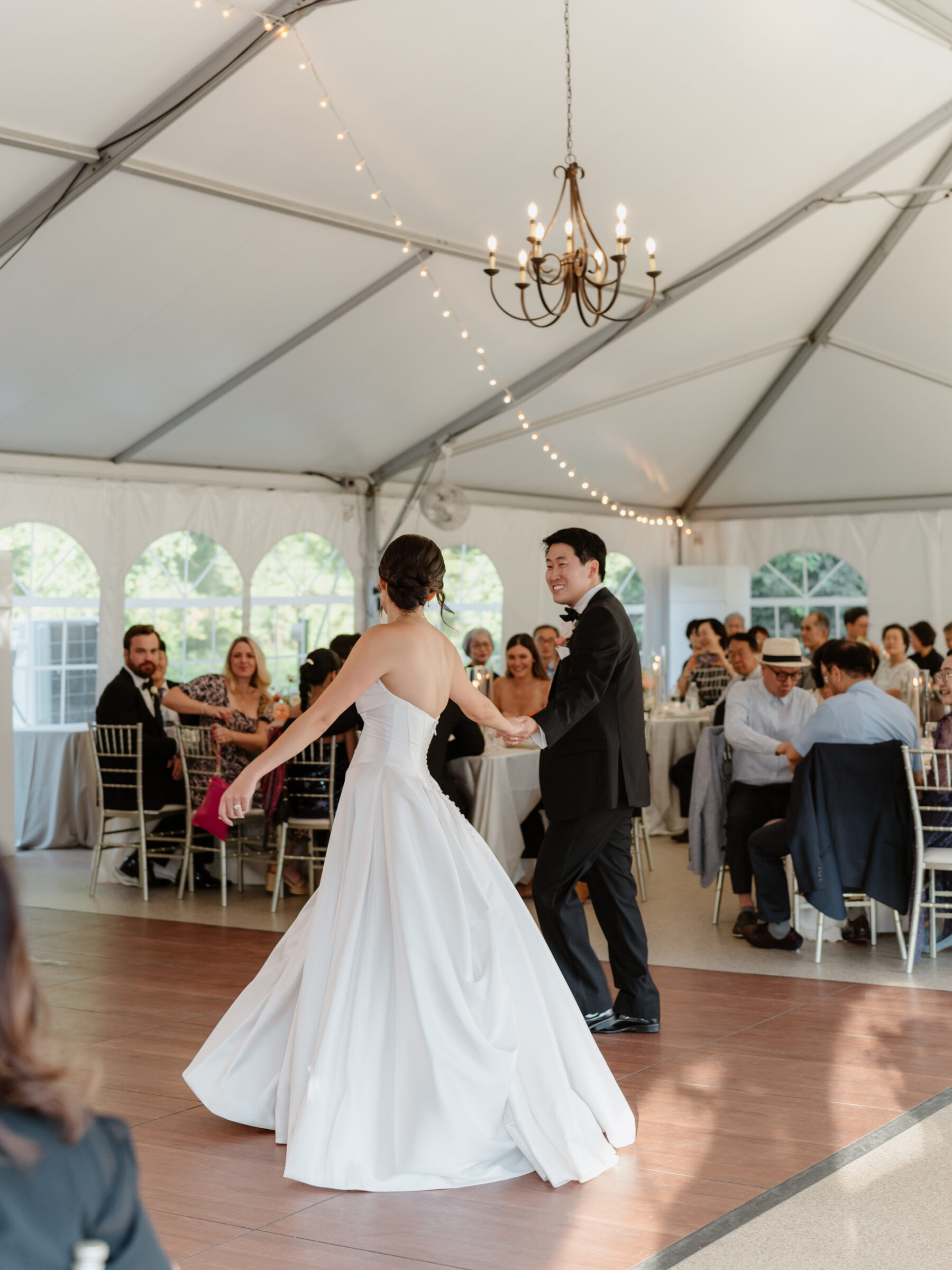 Bride and groom sharing their first dance inside the tented reception, with guests smiling and watching as the bride’s gown twirls across the dance floor.