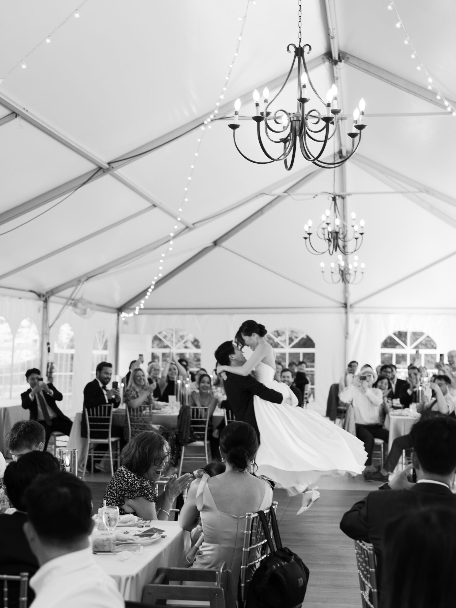 Black and white photo of the groom lifting the bride during their first dance, surrounded by cheering guests inside the tented reception at Rust Manor House in Leesburg VA.