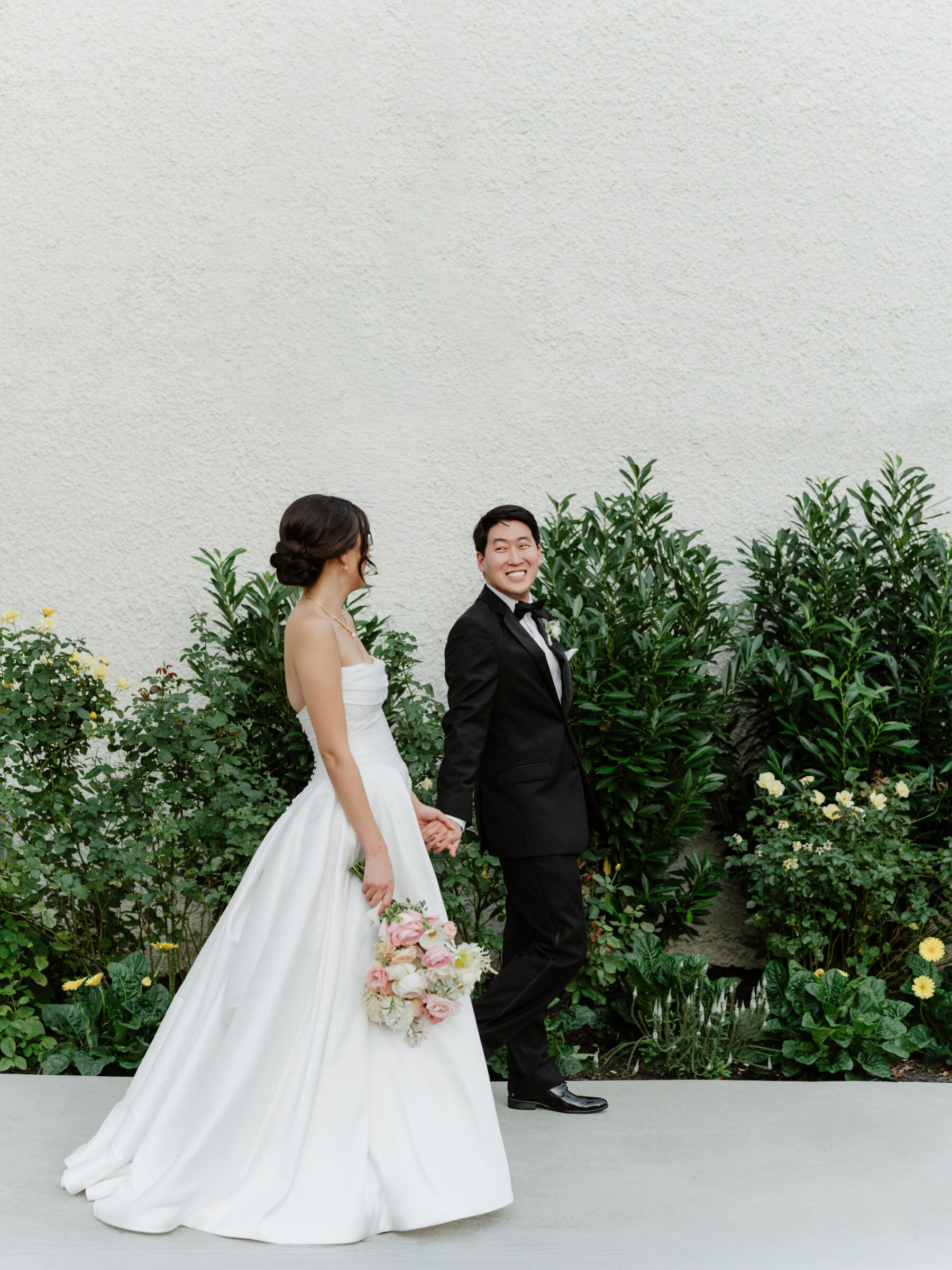 Bride and groom walking hand in hand along the garden pathway, with the bride’s bouquet and gown flowing softly as they share a joyful moment.