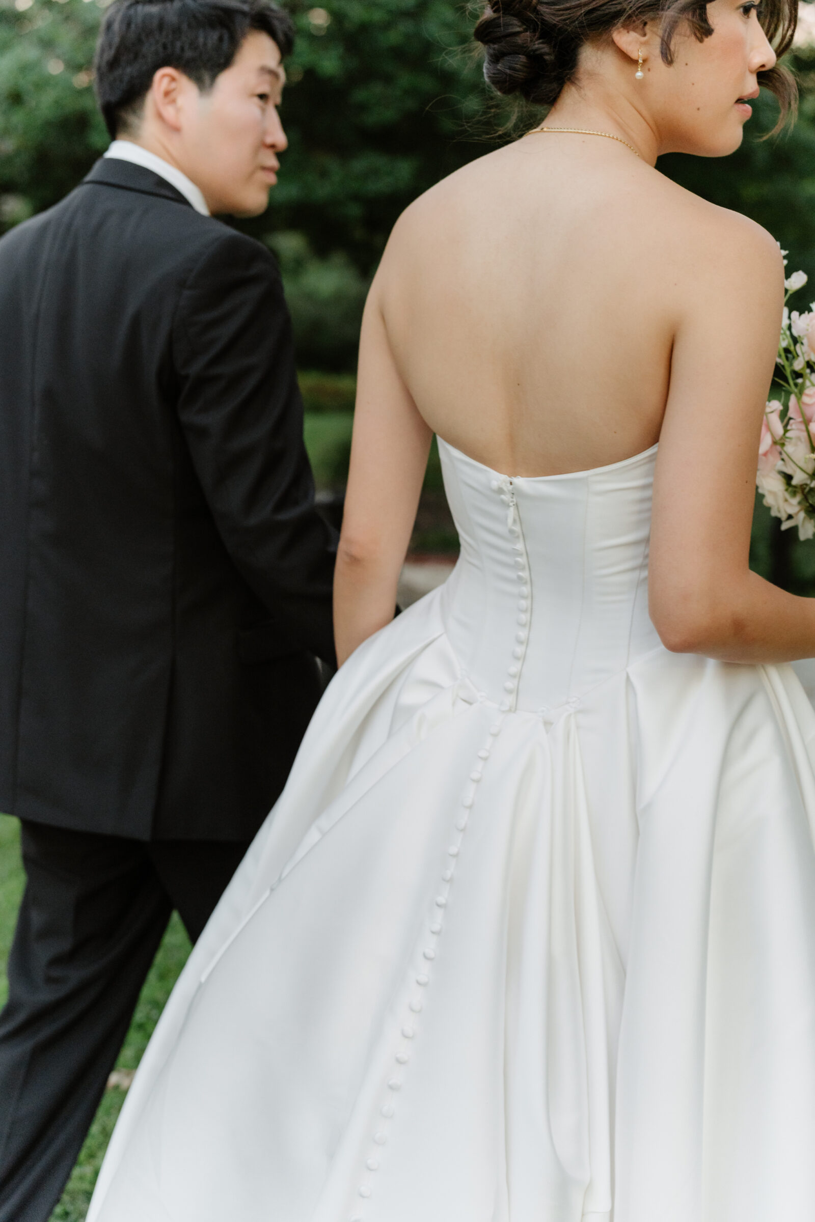 Close-up of the bride’s gown and buttoned back as she walks hand in hand with the groom through the garden.