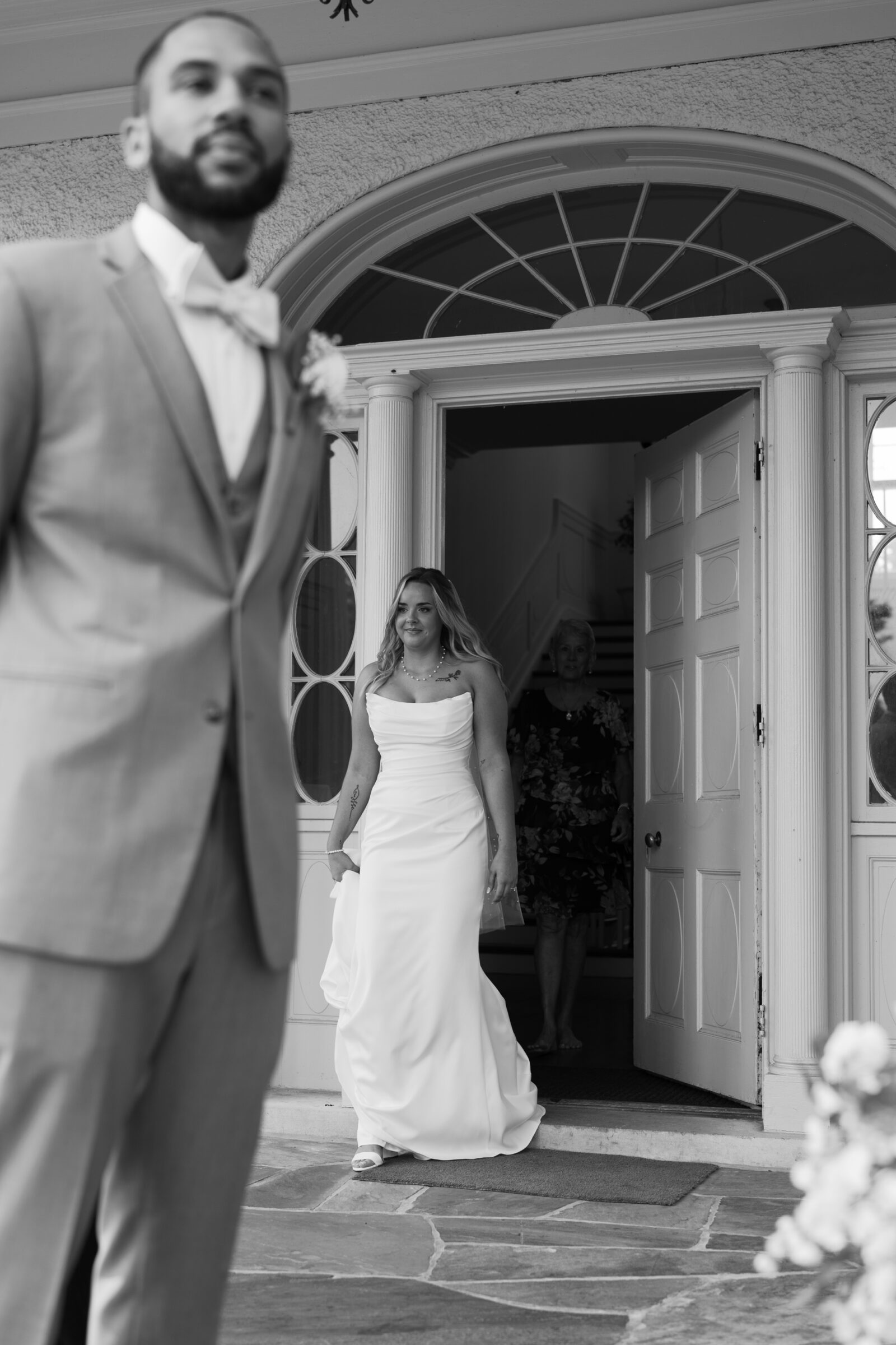 Bride walking out of the historic front entrance at Rust Manor House as the groom waits for their first look, captured in black and white.