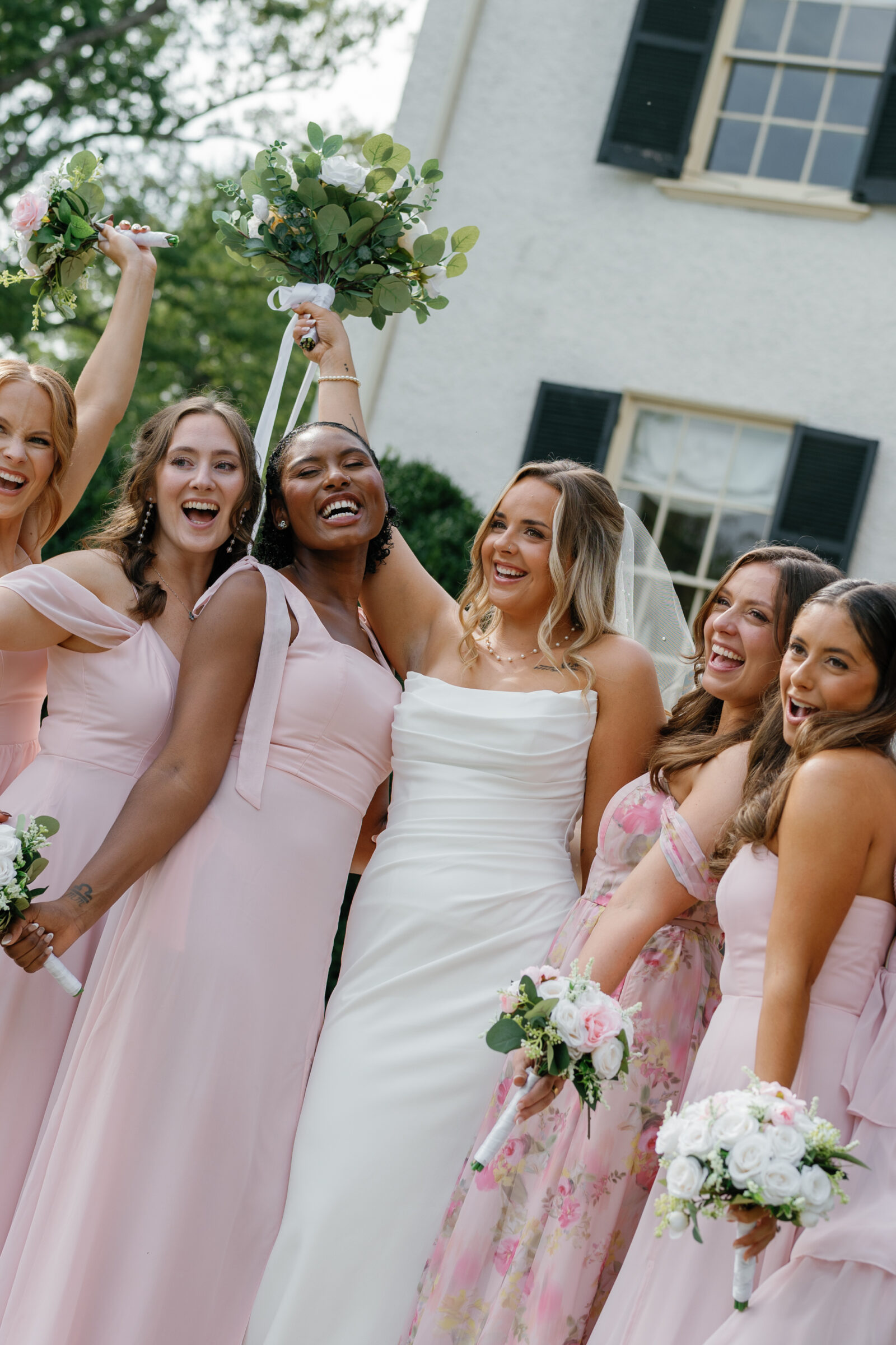 The bride surrounded by her bridesmaids in pink and floral gowns, holding bouquets and celebrating outside Rust Manor House.