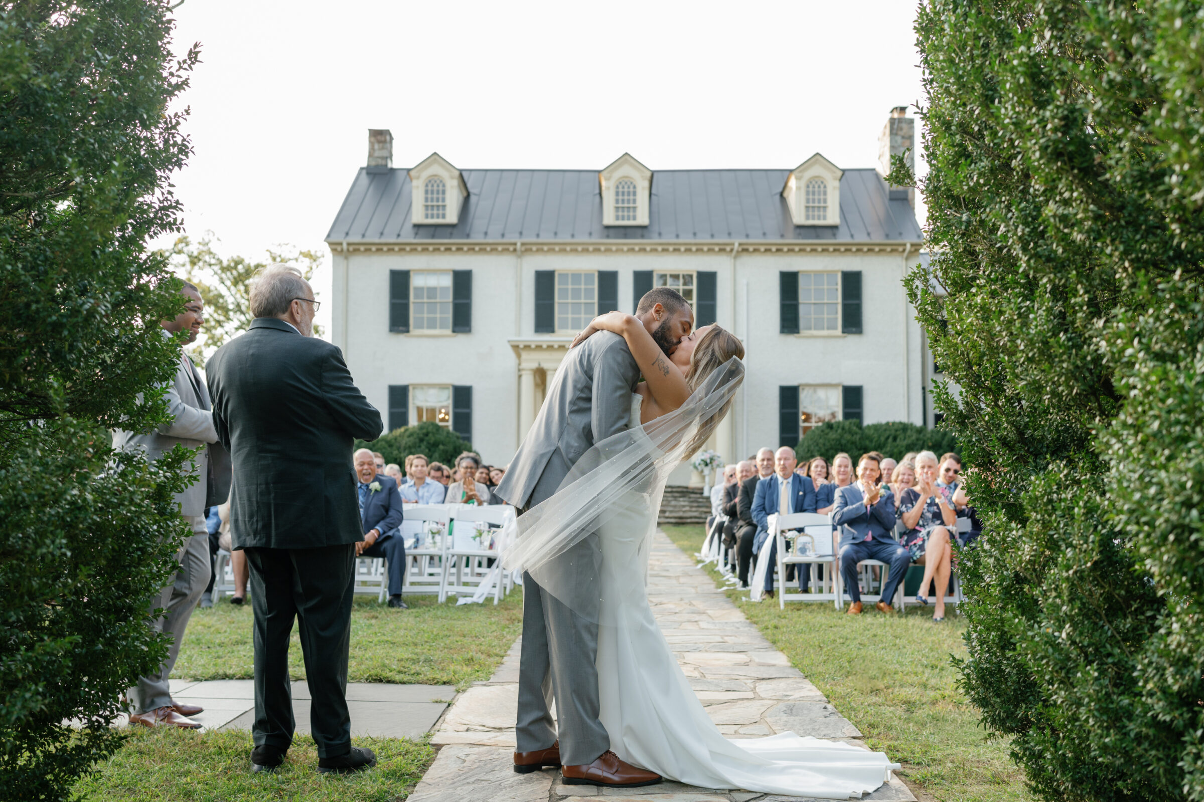 The bride and groom share their first kiss during their outdoor ceremony at Rust Manor House as guests cheer behind them.