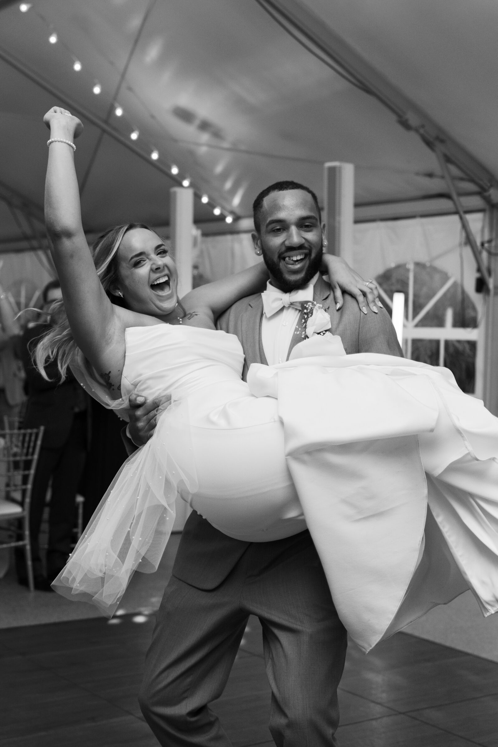 The groom lifts the bride on the dance floor during their reception, both smiling and celebrating under the tent lights.
