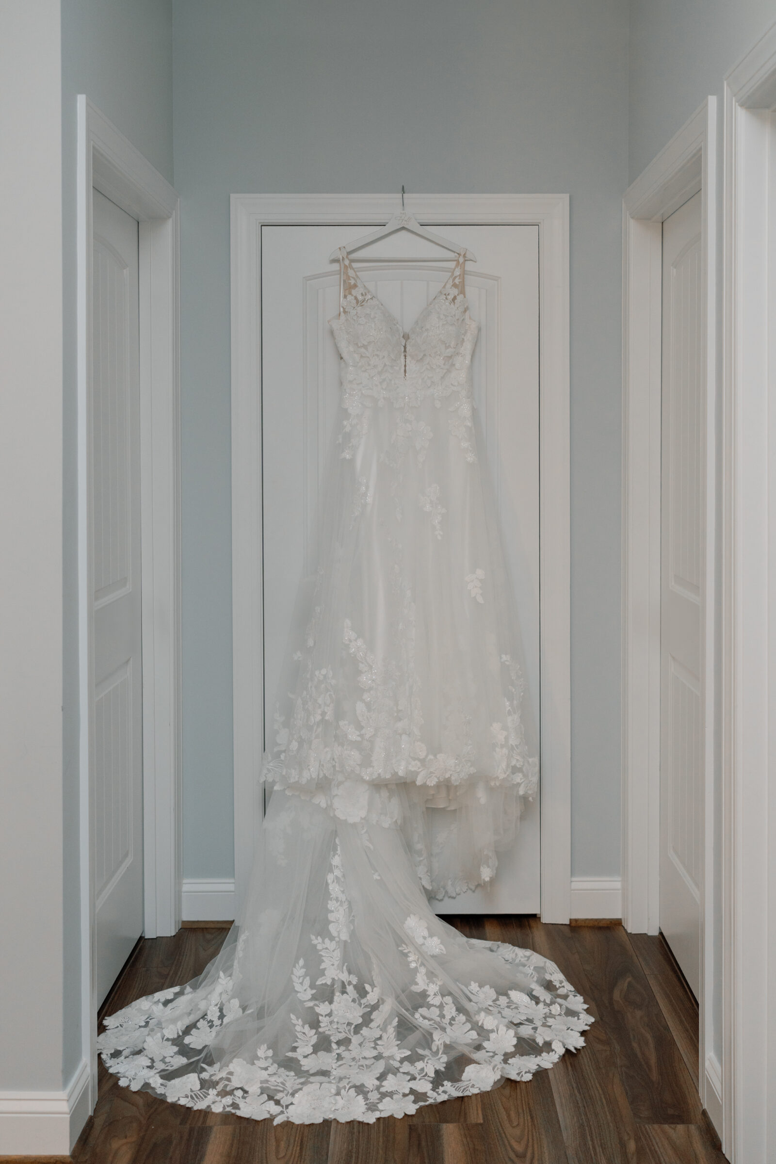 Wedding dress with floral lace detailing hanging in a softly lit hallway, showcasing its delicate train and romantic spring texture.