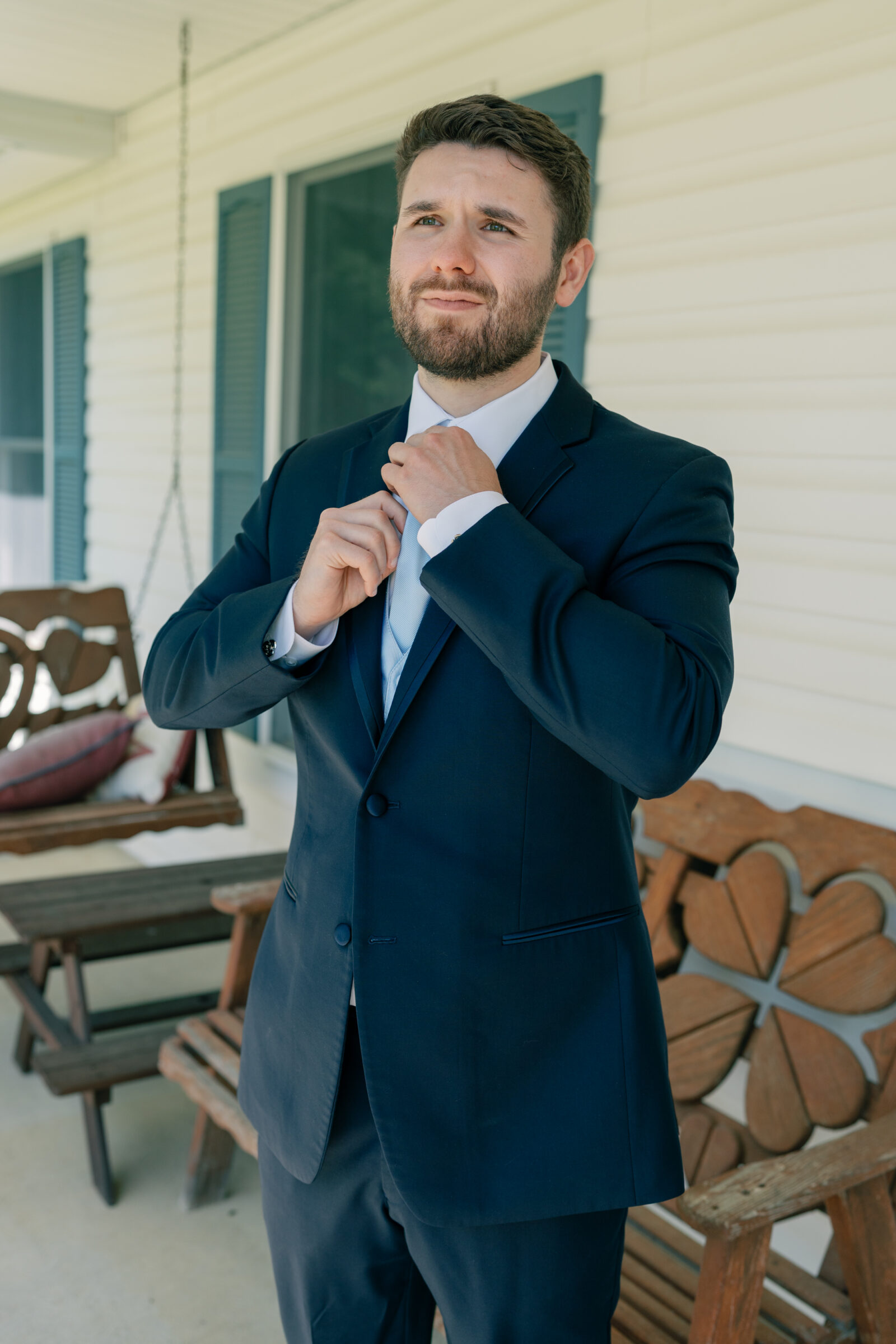 Groom adjusting his tie on a front porch, dressed in a navy suit and captured in natural light for a classic wedding morning portrait.
