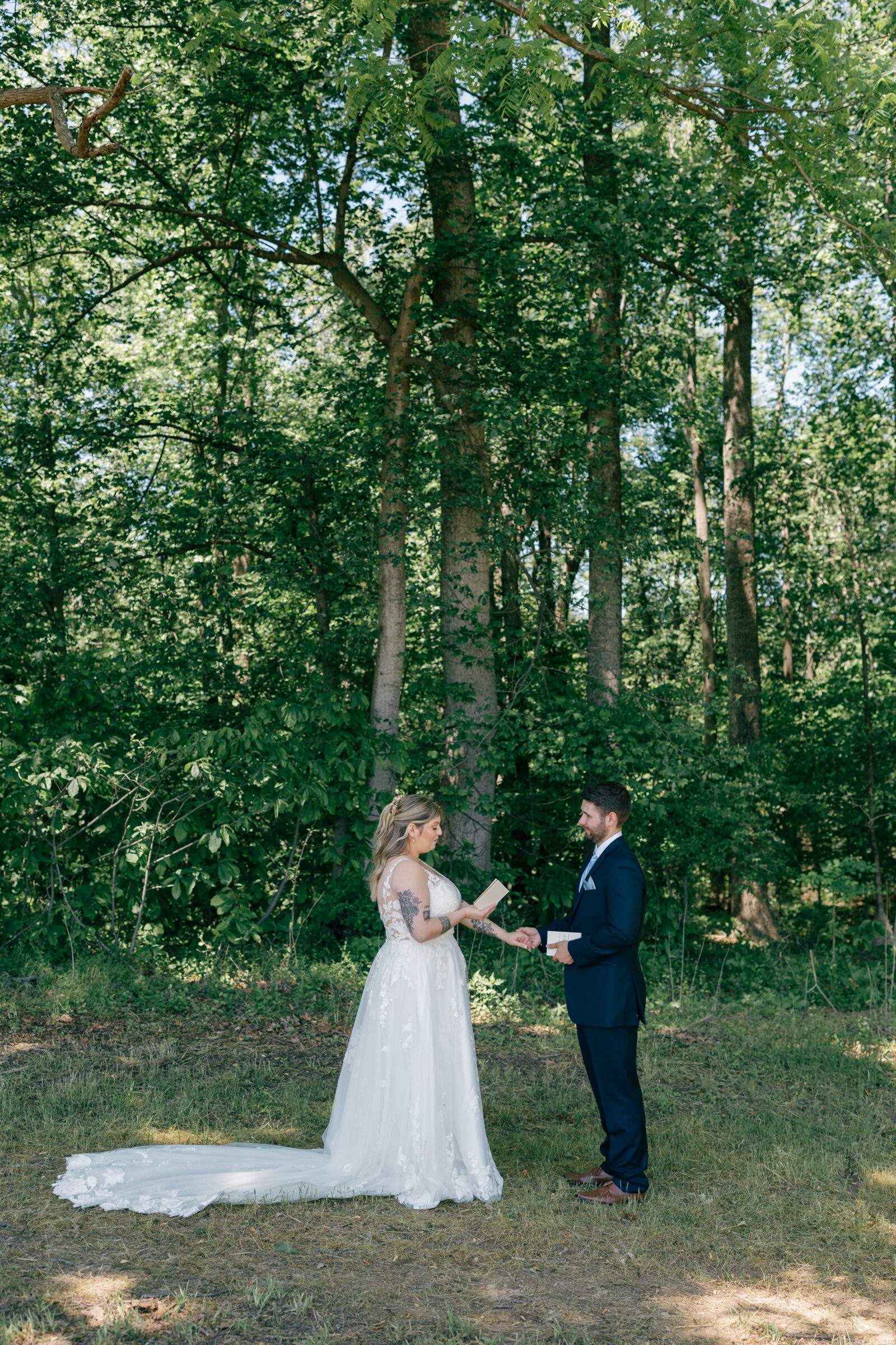 Wide shot of the bride and groom exchanging vows in a sunlit forest clearing, surrounded by tall trees and soft summer greenery.