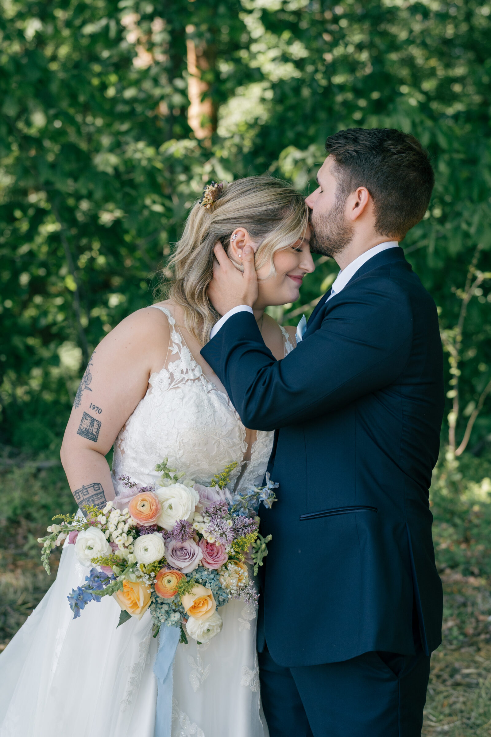 Southern Maryland Wedding Photographer | Groom kissing the bride’s forehead as she smiles softly, holding a colorful wildflower bouquet against a backdrop of lush summer greenery.