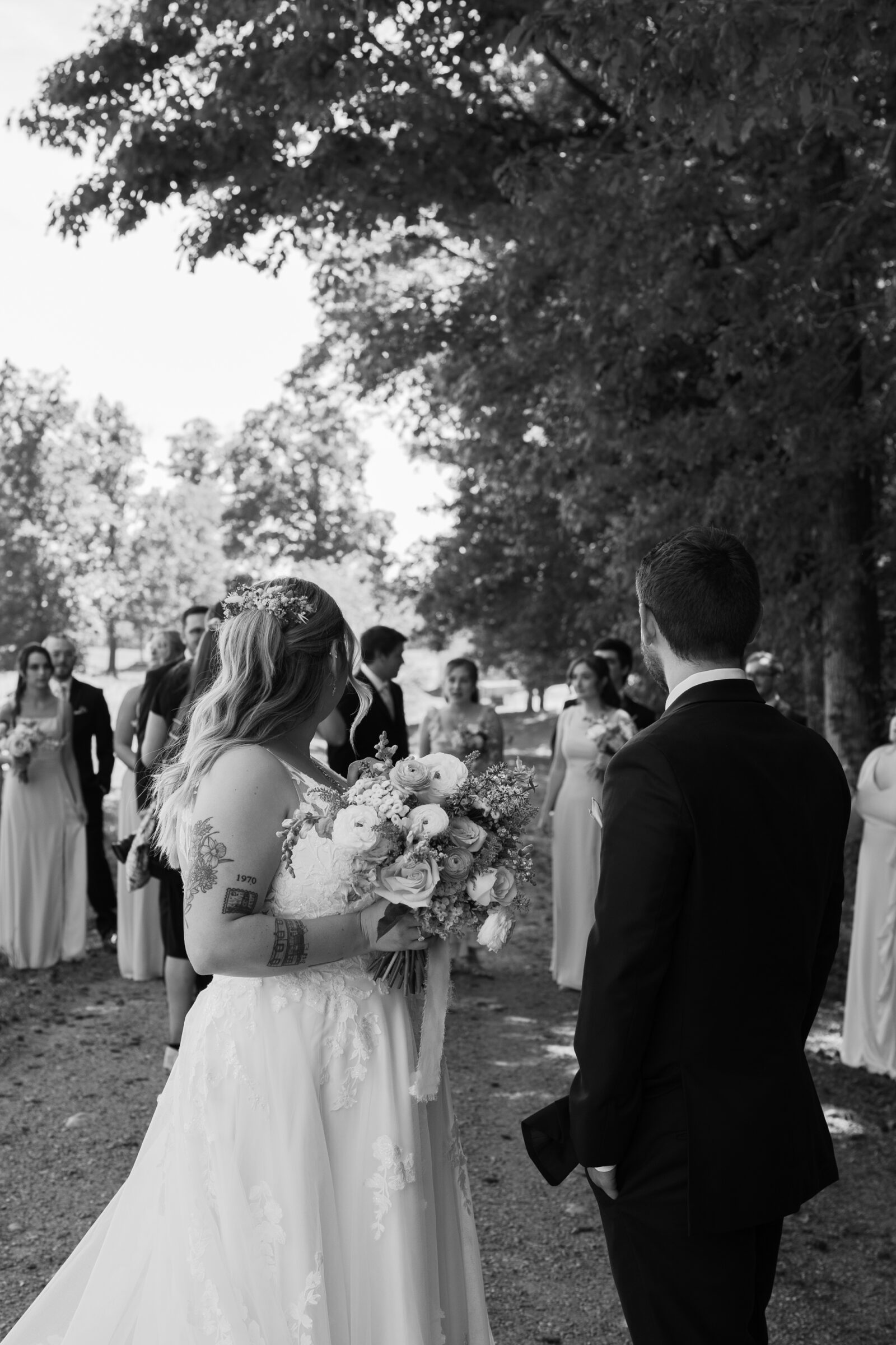 Southern MD Wedding Photographer | Black and white candid of the bride and groom walking toward their wedding party, the bride holding her bouquet as the group gathers under shaded trees.