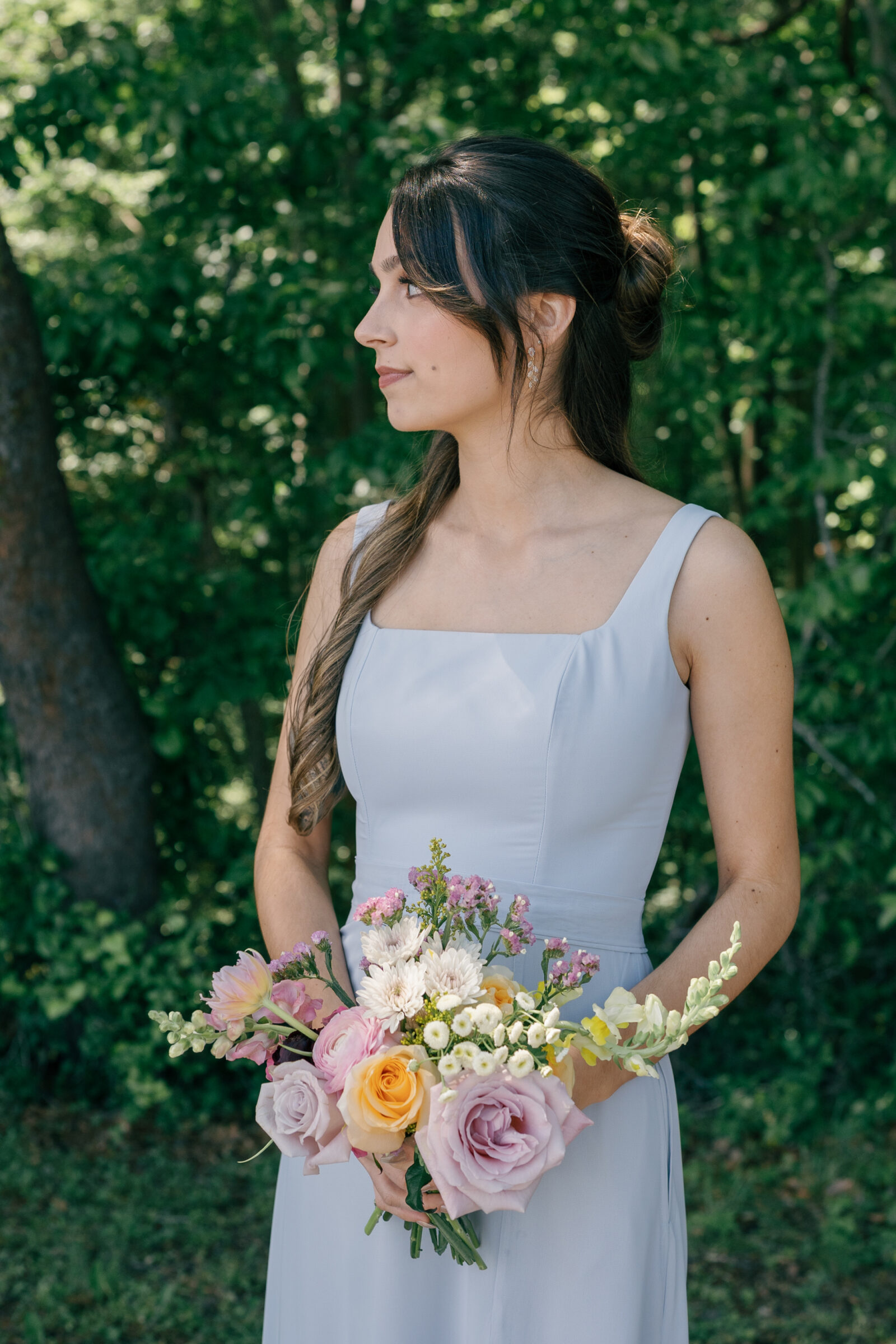 Southern Maryland Wedding Photographer | Bridesmaid in a light blue dress holding a pastel wildflower bouquet, standing in soft natural light against a wooded backdrop.