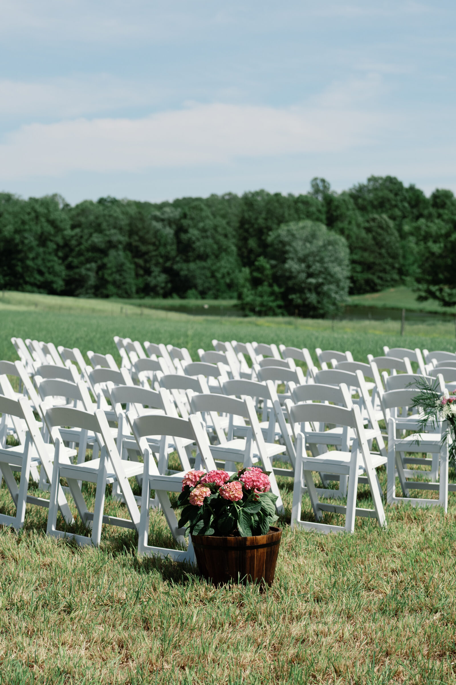 Rows of white ceremony chairs set up in an open field, with a pot of pink hydrangeas adding a soft floral touch against the countryside backdrop.