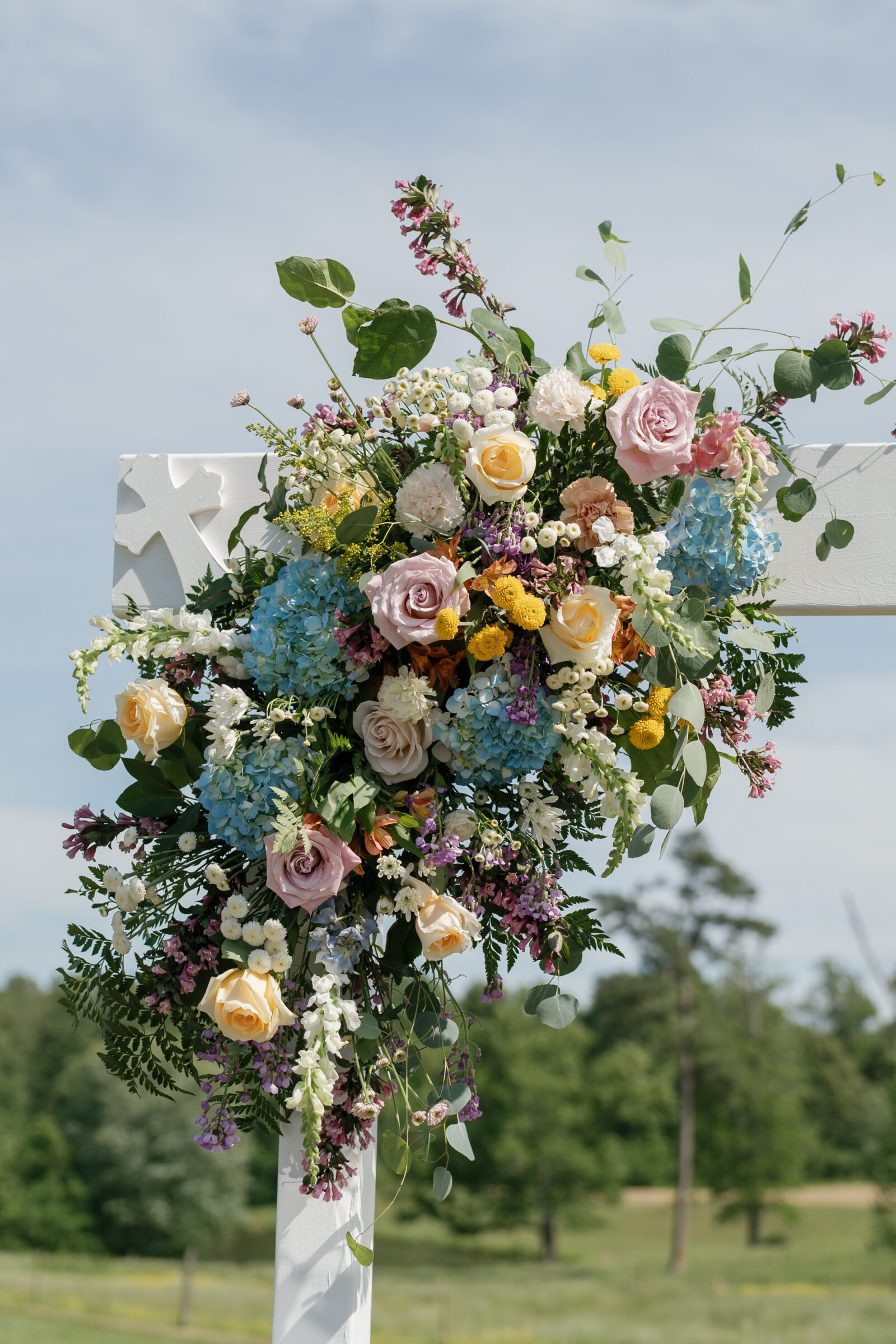 Colorful floral arrangement of roses, hydrangeas, and wildflower-inspired blooms decorating a white ceremony arch against an open field backdrop.