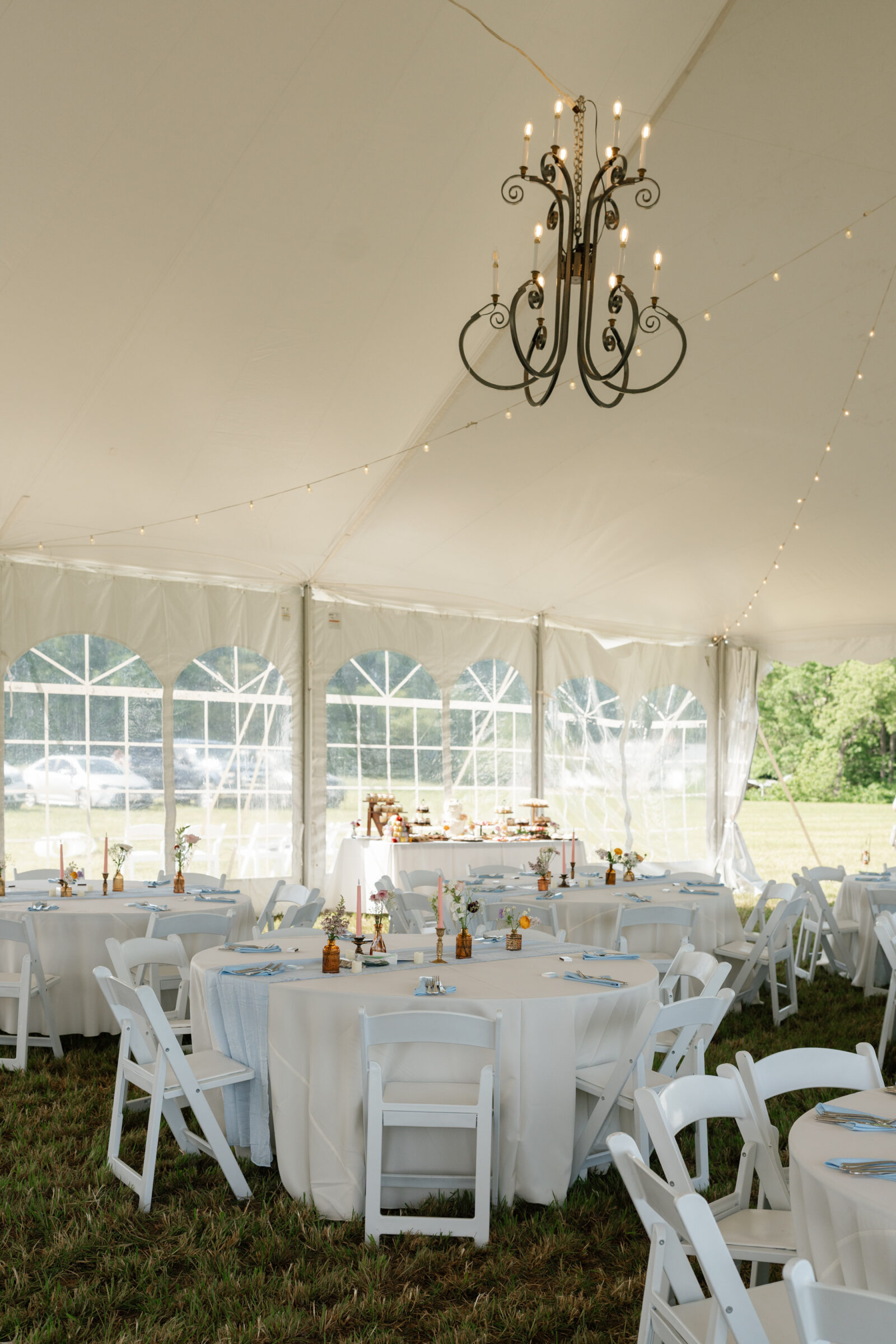 Tented wedding reception with round tables, white chairs, pastel candles, and delicate floral centerpieces arranged beneath a chandelier and string lights.