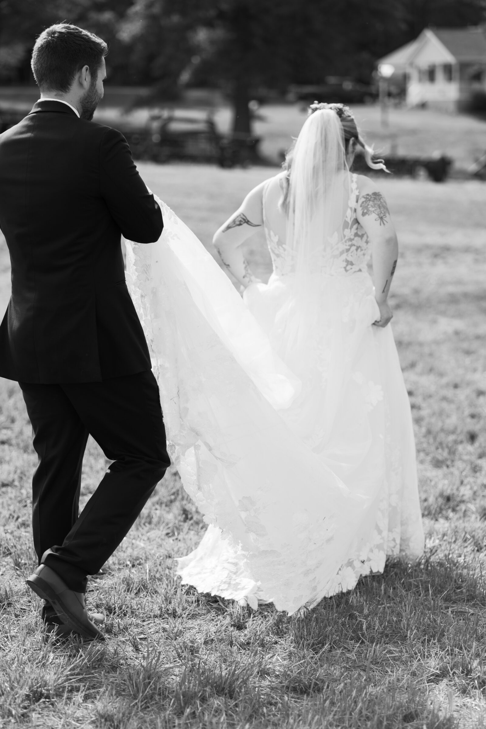 Southern MD Wedding Photographer | The groom carries the bride’s train as the couple walks together across the grass after their ceremony.