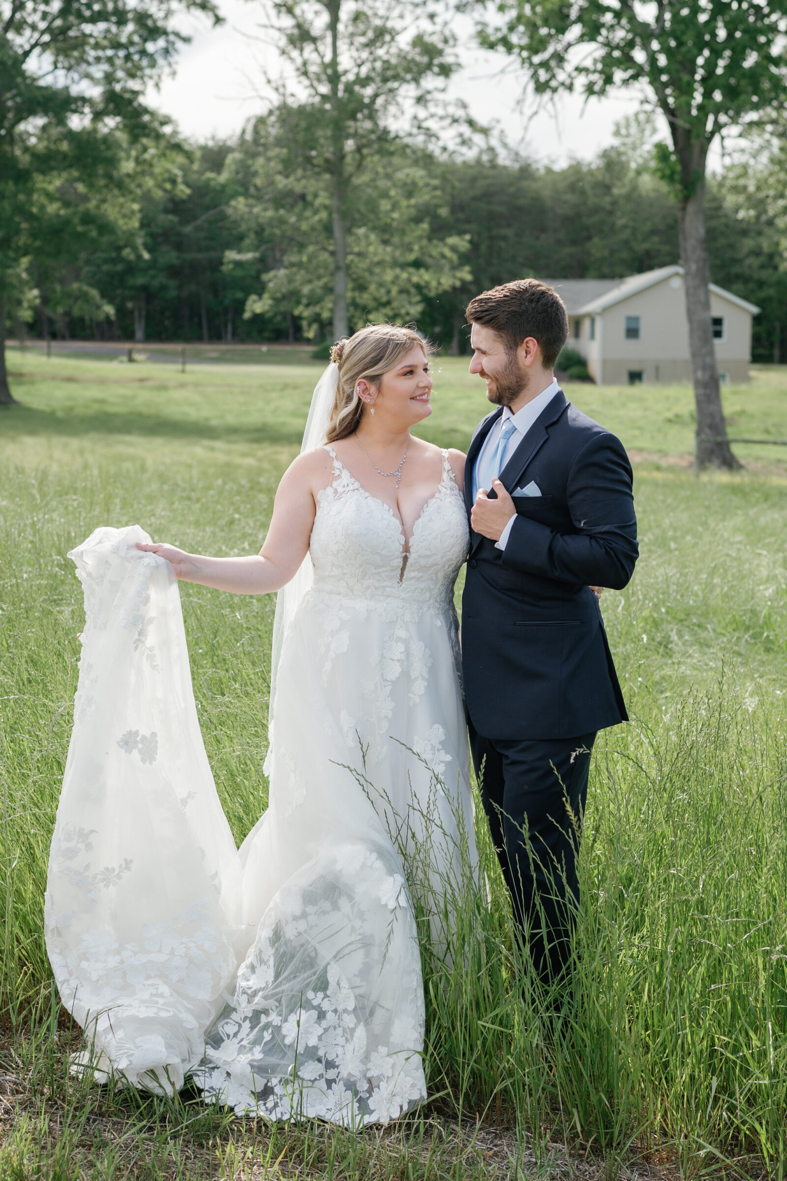 The bride lifts part of her dress as she smiles at the groom while standing together in tall grass.