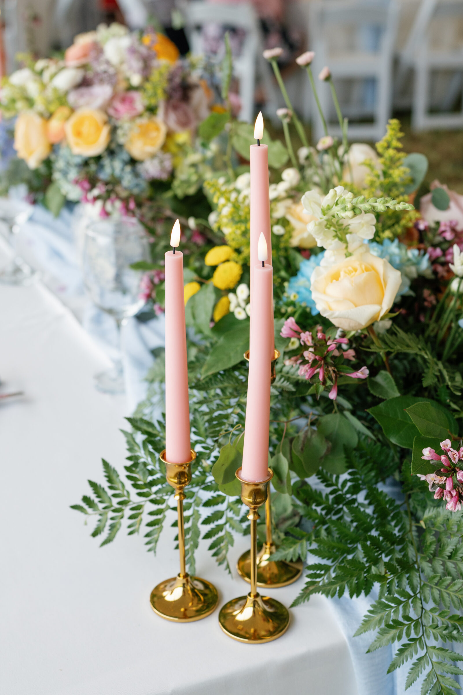 Pink taper candles in gold holders sit beside a lush floral arrangement on a wedding reception table.